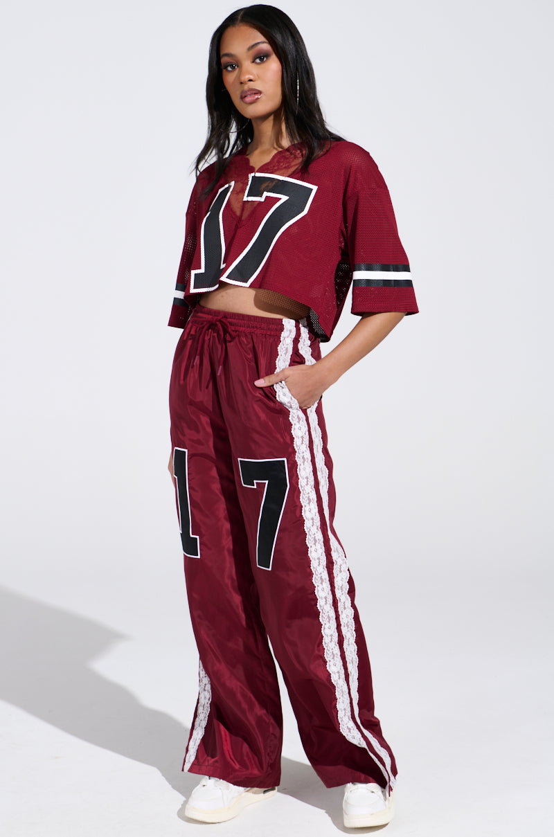 Wearing the sporty-chic LETS GET IT JERSEY— a maroon mesh cropped top with wide-leg pants featuring the number 17—this person pairs the look with white sneakers against a plain backdrop.