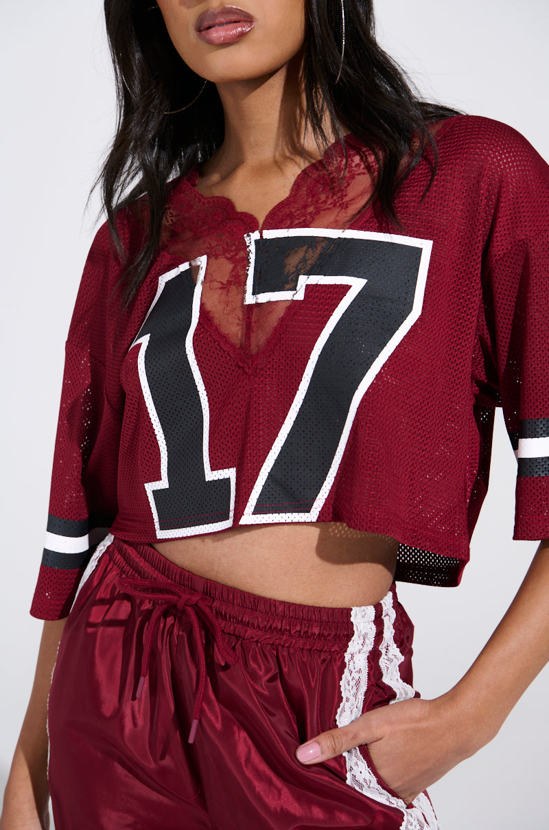 Someone wears the LETS GET IT JERSEY, a maroon sporty-chic top with a bold white and black number 17, paired with matching maroon track pants featuring lace trim, blending athletic flair with fashion-forward style.