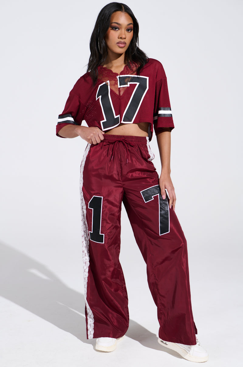 A woman confidently poses in a red sports-inspired outfit featuring a cropped jersey with the number 17 and LETS GET IT TRACK PANT, both in windbreaker fabric. The high-rise relaxed fit pants complement her white sneakers as she stands against a white background.