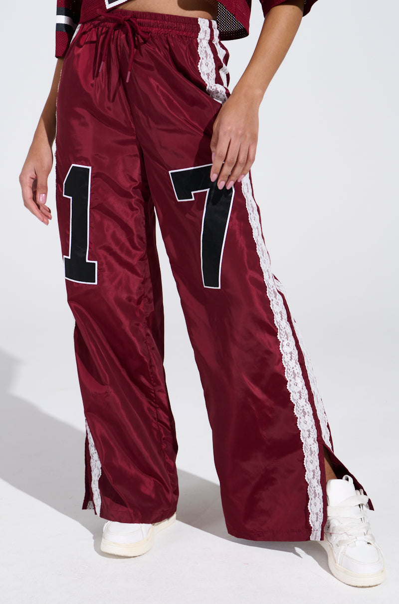 A person is wearing the LETS GET IT TRACK PANT, maroon wide-leg pants with a large "17" on the front, inspired by 90s sweats. They feature white side lace and are paired with white sneakers and a red and white top to complete the high-rise relaxed fit look.