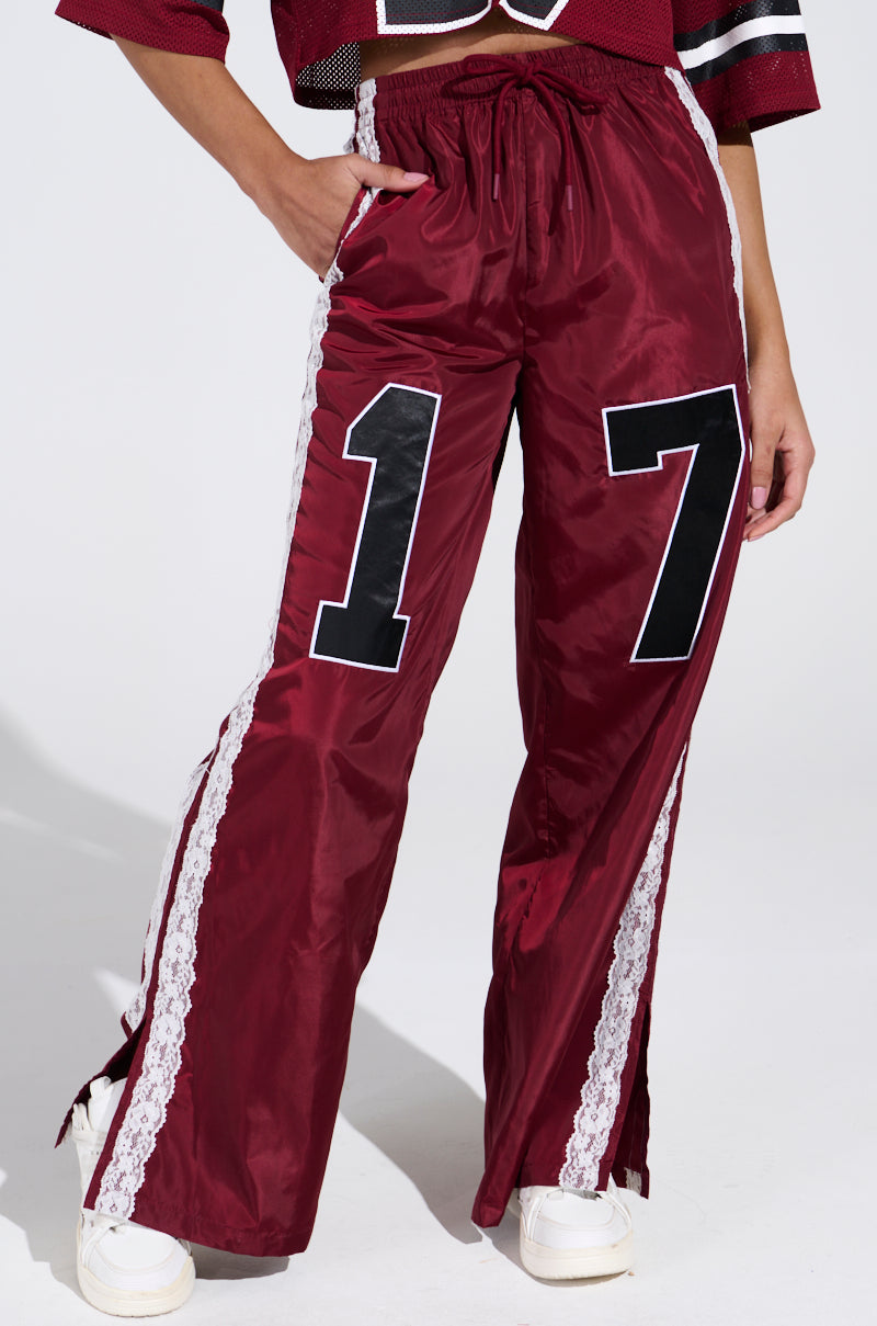 The individual is wearing the LETS GET IT TRACK PANT in maroon, featuring a bold "17" on one leg and white lace trim along the sides. These 90's-inspired sweats boast a high-rise relaxed fit, paired with a partially visible red cropped top.
