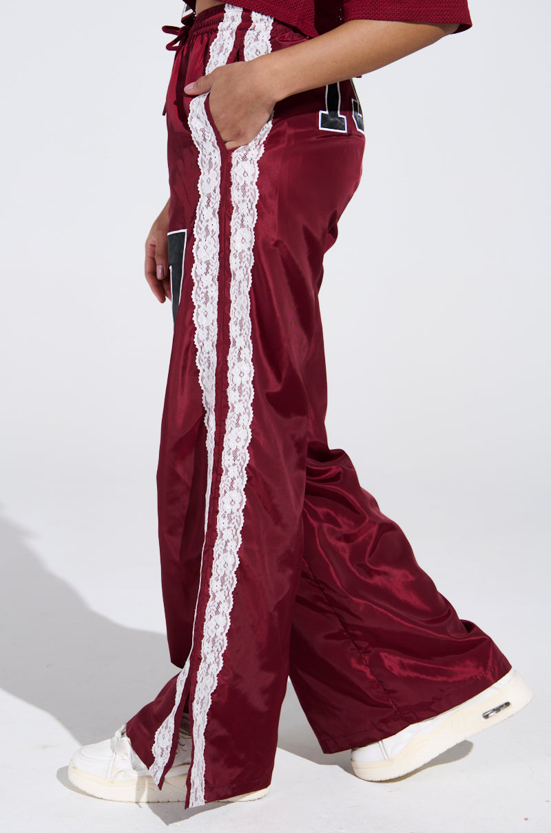 The person is rocking the LETS GET IT TRACK PANT in maroon, featuring a high-rise relaxed fit and white lace stripes akin to 90's sweats. A matching maroon top completes the ensemble while white sneakers pop against the minimalist backdrop.