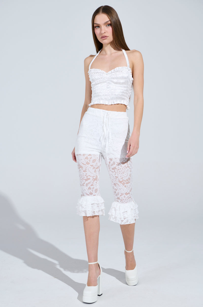A woman with long brown hair elegantly wears a white sleeveless top paired with the GIRLY POP LACE CAPRI LEGGING featuring ruffled hems. She completes her high-rise look with chic white platform heels against a plain, light background. 0