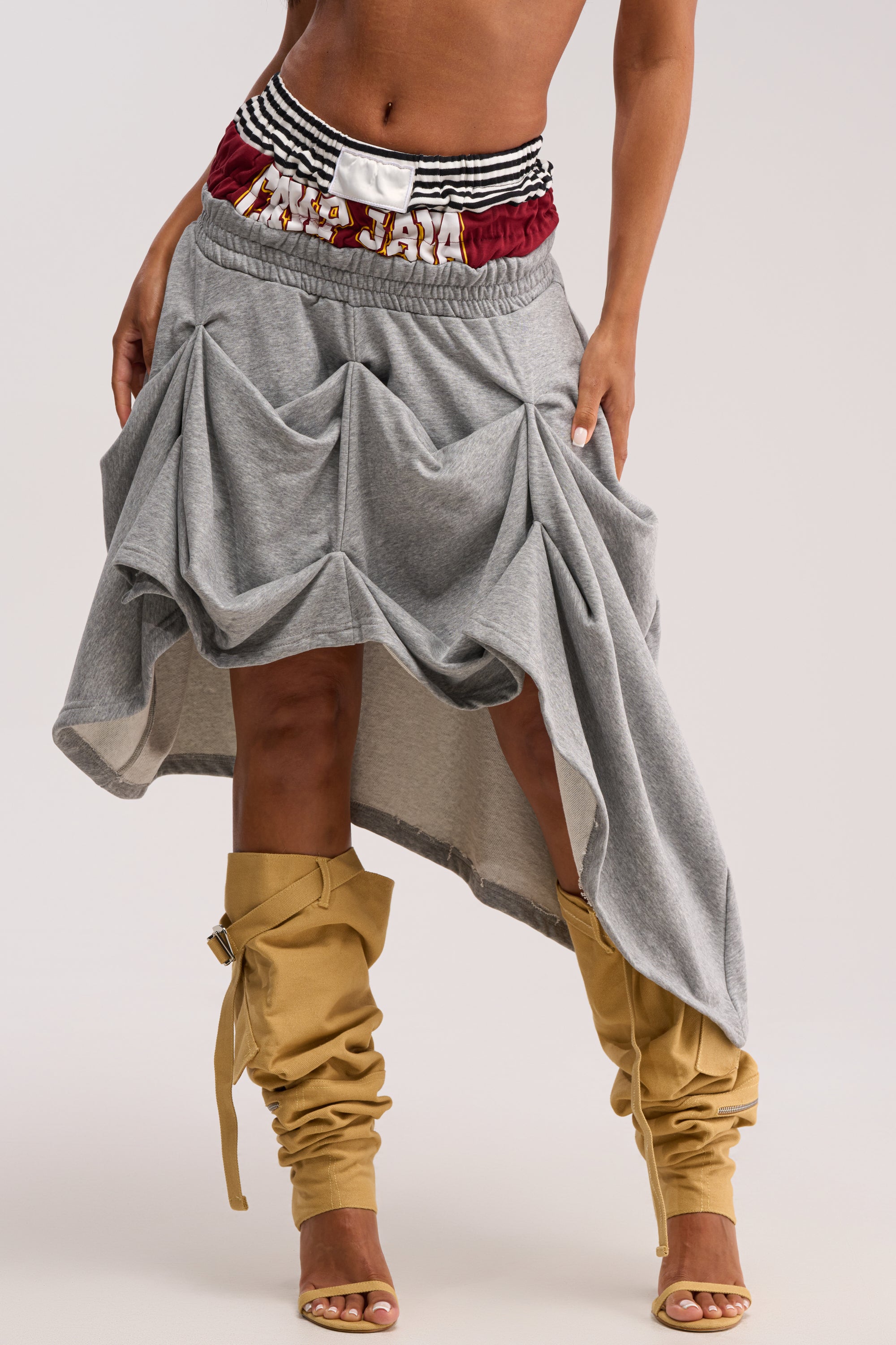 A person wearing the NO REGRETS HERE MIDI SKIRT, a gray sporty ruched skirt with an asymmetrical hem and striped waistband, pairs it with tall mustard-yellow slouchy boots against a plain background.