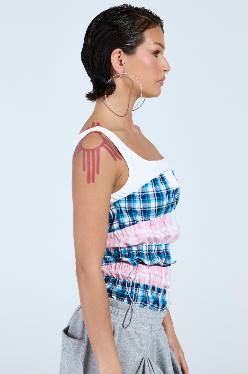 A person with short, dark hair and large hoop earrings wears the DONT MESS WITH ME TANK in a vibrant plaid pattern, paired with grey pants. They have a stylized red dripping tattoo on their upper arm against a light blue background.