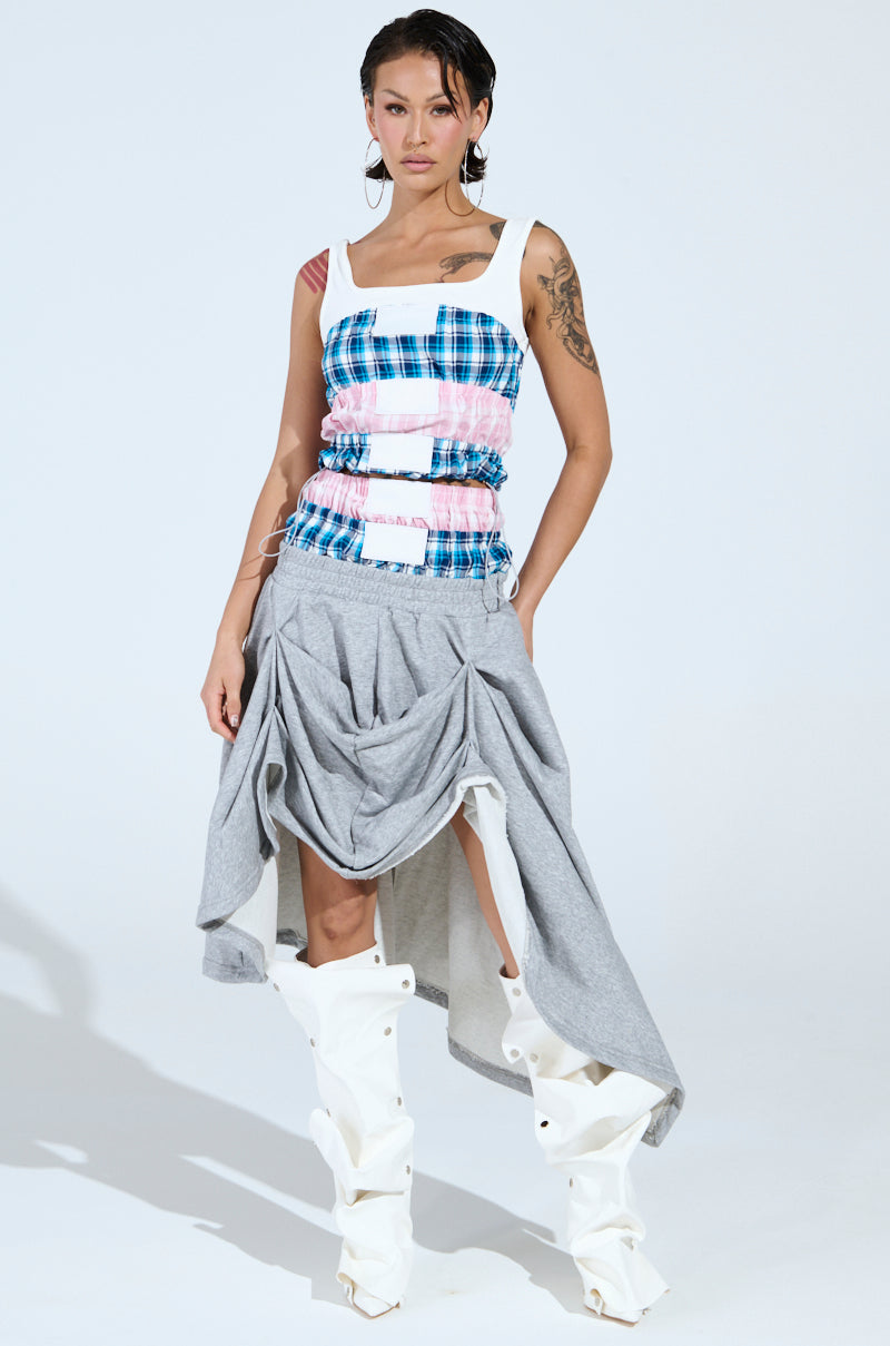 A woman faces forward, wearing the DONT MESS WITH ME TANK with a colorful plaid pattern, a gray draped skirt, and tall white sculptural boots. She has tattoos on her arms and slicked-back wet-look hair.
