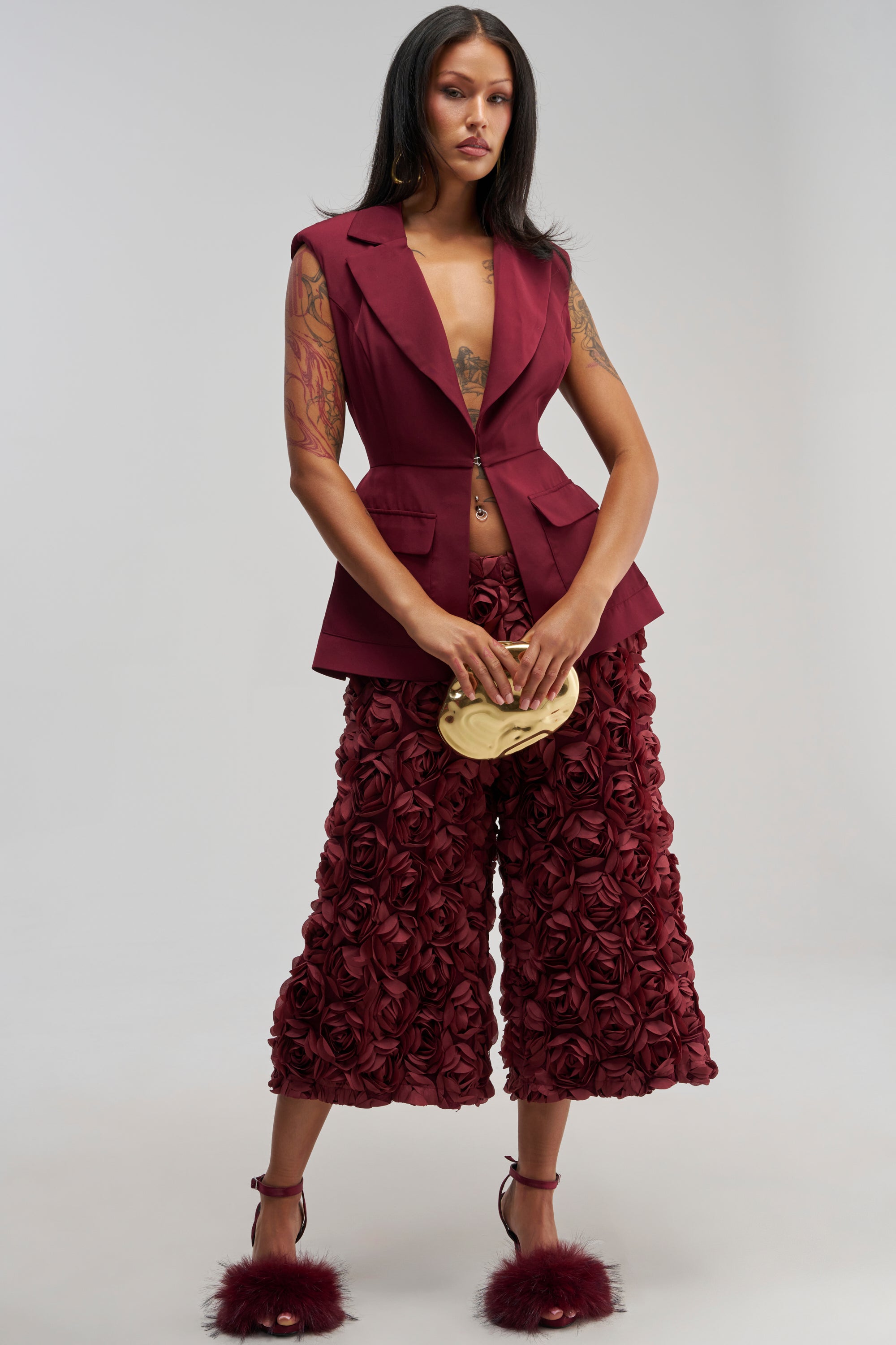 A woman wears the ROSE PETAL GAUCHO burgundy floral pants, styled with a sleeveless maroon blazer, maroon feathered heels, and a gold clutch as she stands against a plain background.