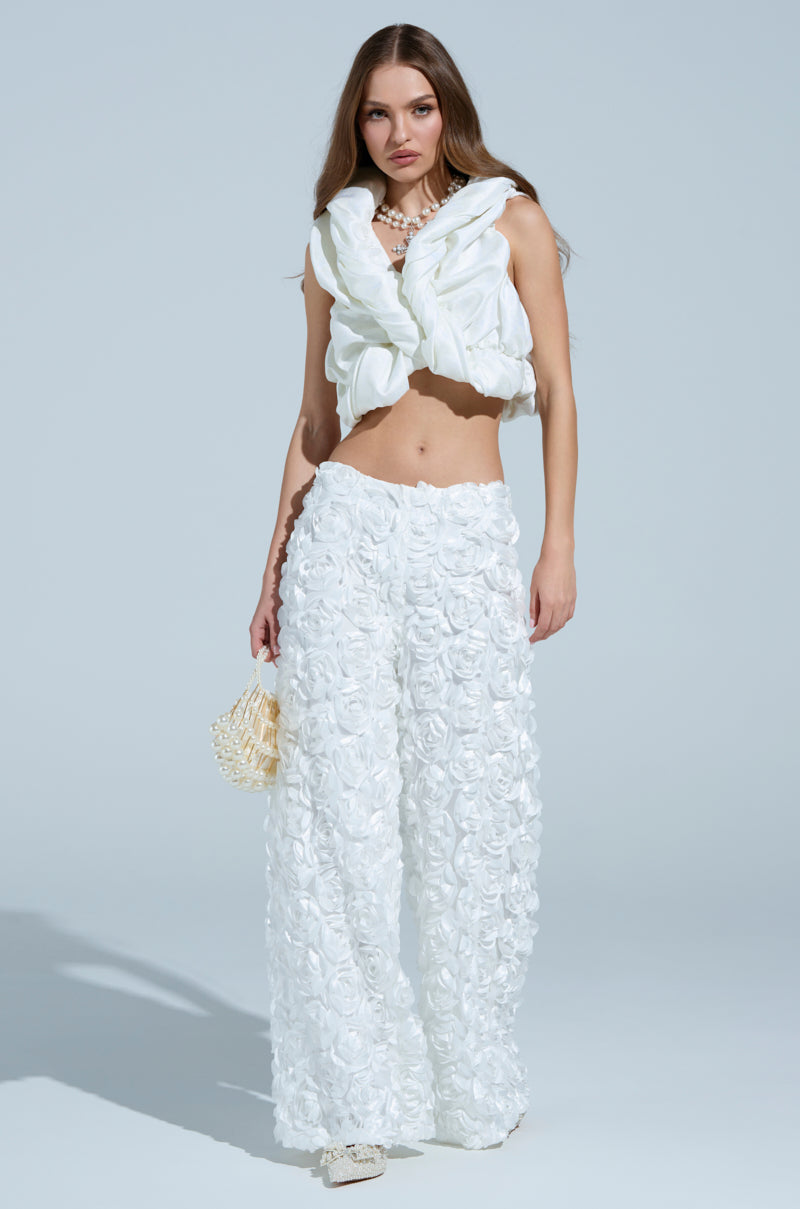A woman poses stylishly in a sleeveless white top with ruffles and ROSE PETAL WIDE LEG PANTS featuring 3D rose appliques. She carries a small woven bag, wears white shoes, and stands against a light blue background.
