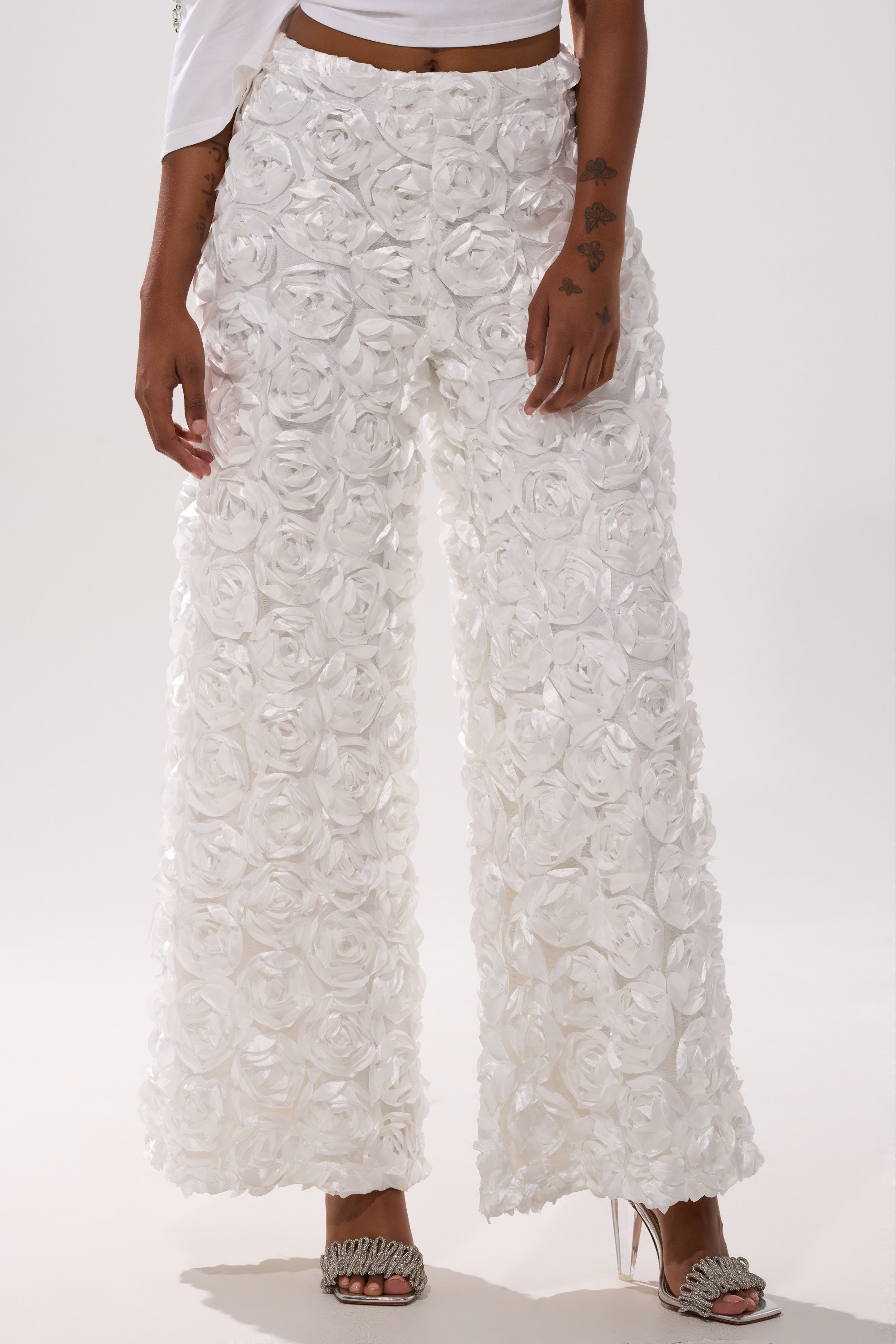 A person wears the ROSE PETAL WIDE LEG PANT, featuring 3D floral rosettes, with a cropped white top and open-toe heeled sandals against a plain light background.