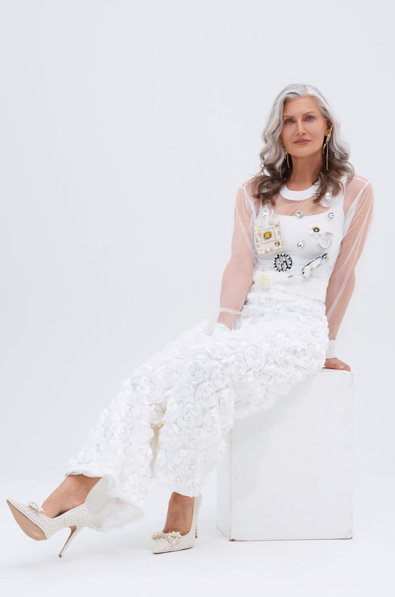 A woman with long gray hair, wearing the ROSE PETAL WIDE LEG PANT and a sheer top, sits on a white block against a plain background. She has crossed her ankles and wears embellished heels.