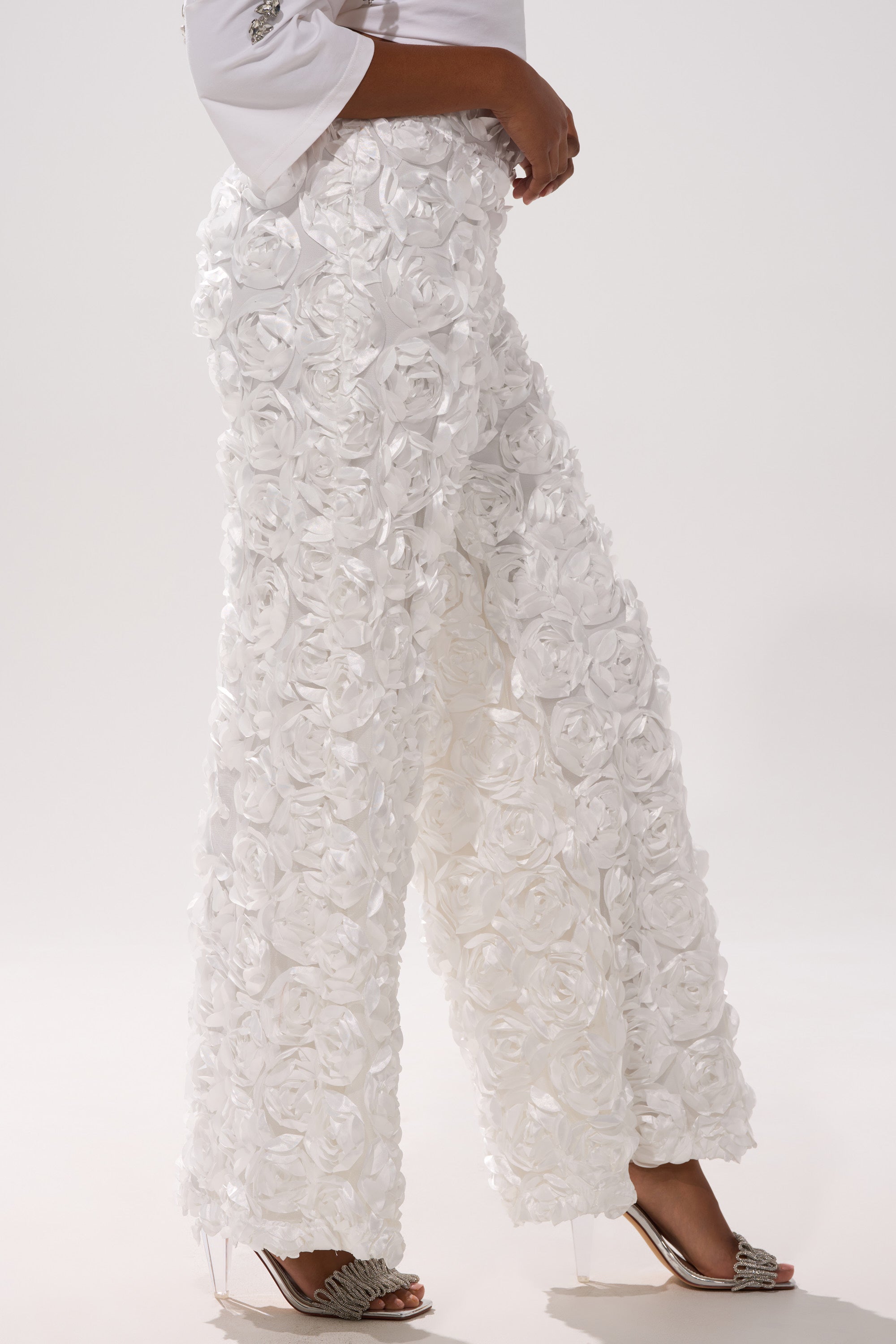Someone wears the ROSE PETAL WIDE LEG PANT, featuring white mesh and 3D satin rose appliques, styled with a white top and open-toe heels against a plain background.