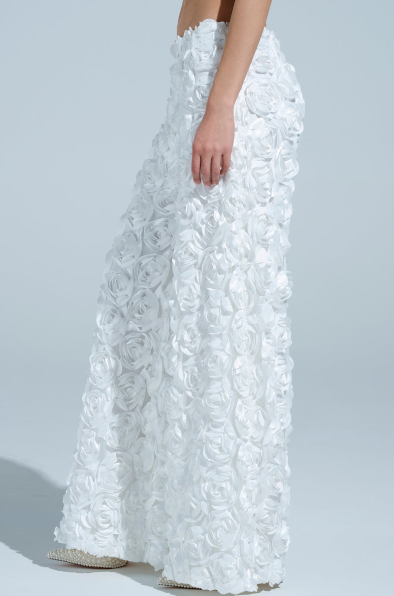 Someone is wearing the ROSE PETAL WIDE LEG PANT, characterized by white fabric with a textured rose pattern. These high-waisted pants fit at the waist and elegantly flare out at the bottom against a plain light blue background.