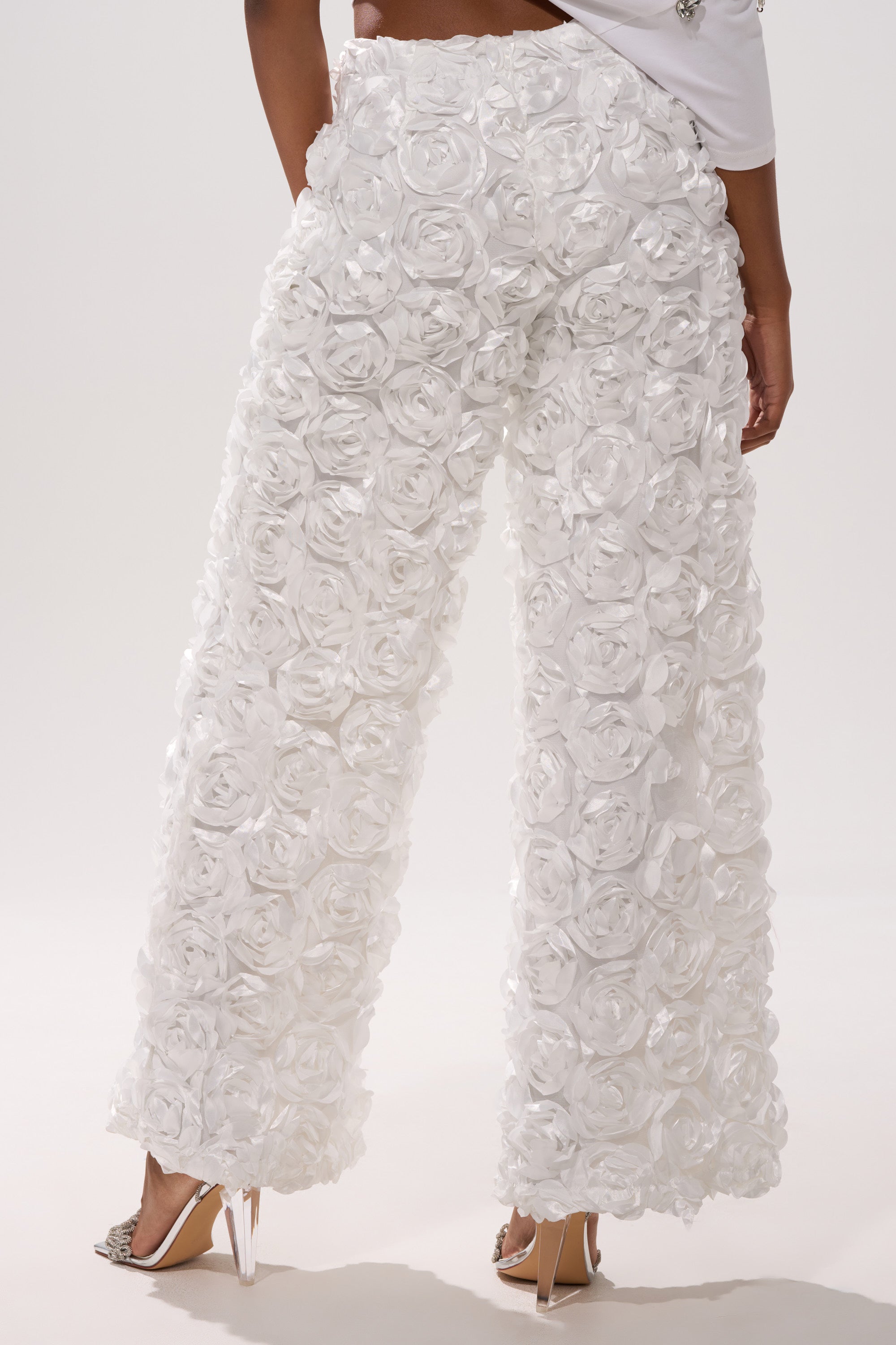 A person in high heels and the ROSE PETAL WIDE LEG PANT, featuring textured 3D satin rose appliques, is seen from the back against a plain light background.