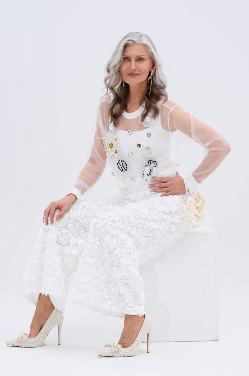 A stylish older woman with gray hair sits on a white block, wearing the ROSE PETAL WIDE LEG PANT, a sheer white top with satin rose appliques, pearl-embellished heels, and gold earrings against a plain white background.