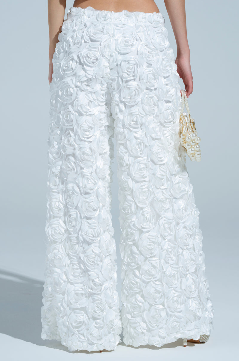 A person wears the ROSE PETAL WIDE LEG PANT featuring textured floral patterns and rose appliques, paired with a small handbag adorned with pearl details. The light gray background highlights the intricate fabric of these mid-rise pants.
