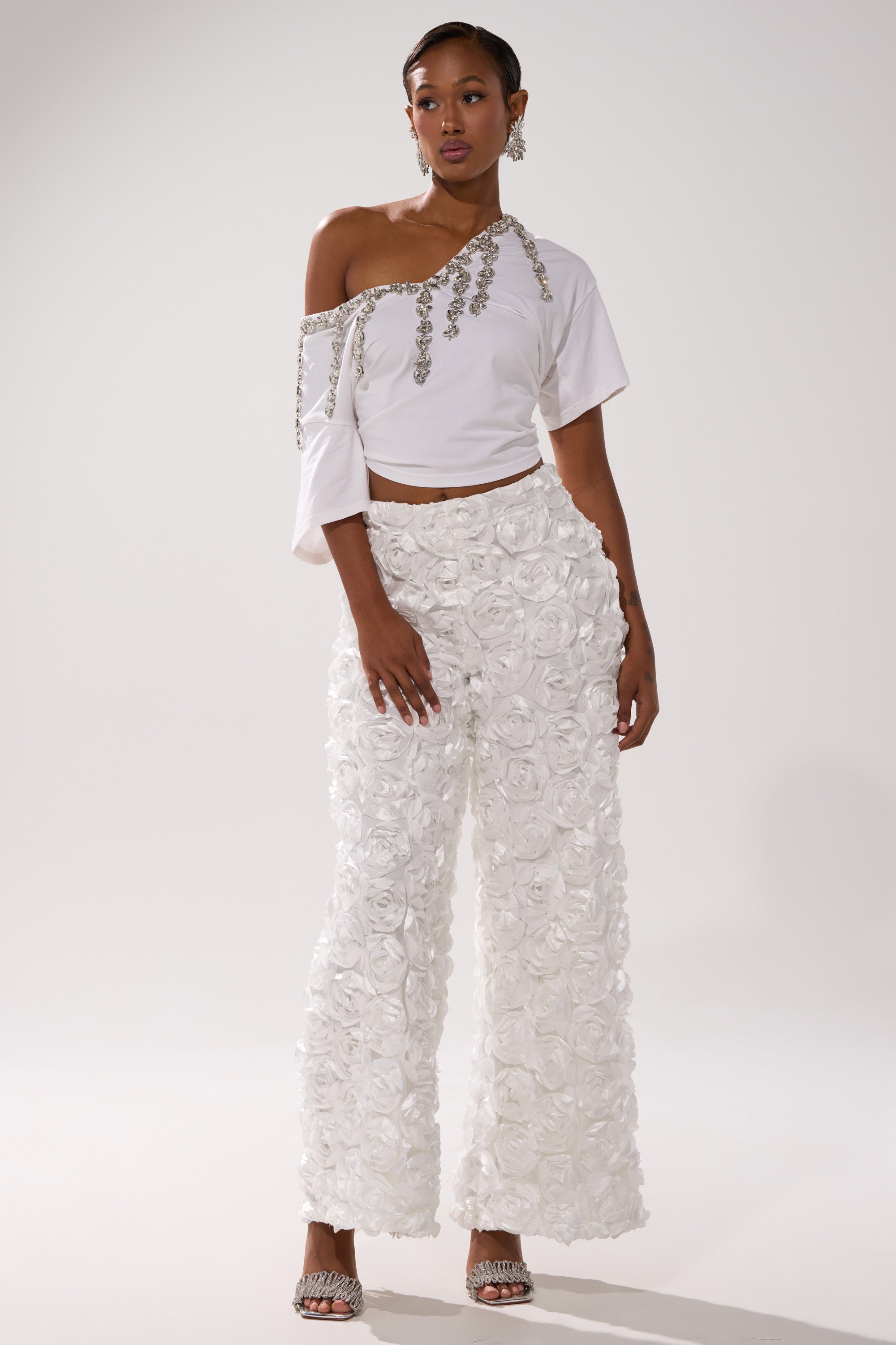 A woman poses in ROSE PETAL WIDE LEG PANT, featuring high-waisted mesh and textured floral appliqués, paired with a white off-shoulder embellished top, silver heels, and earrings against a plain background.