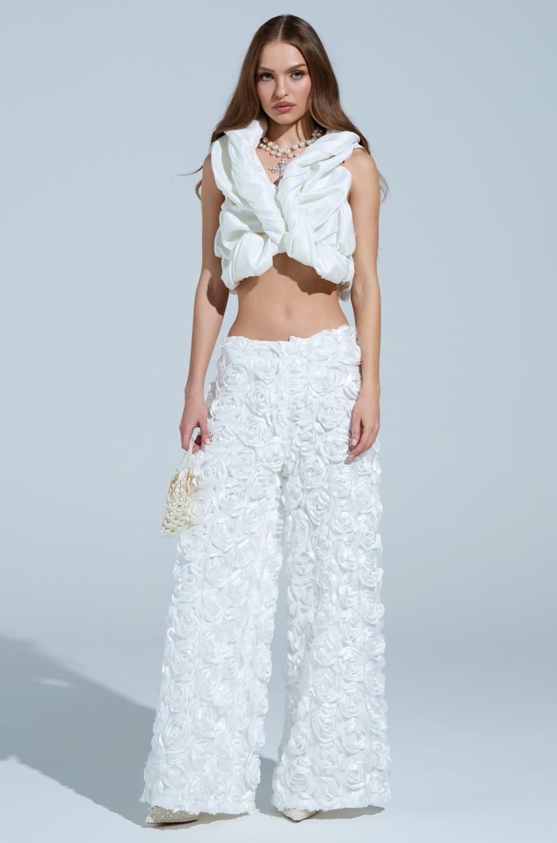 A woman wears a sleeveless white top with a v-neck and ruffles, paired with ROSE PETAL WIDE LEG PANTS featuring textured floral patterns. She carries a small beaded bag and delicate jewelry, all set against a neutral background.
