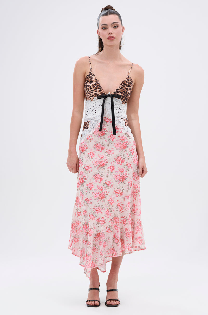 A woman stands against a white background wearing the CARRIE CODED SATIN MIDI DRESS, featuring a leopard print bodice, lace-up front, white lace midsection, and pink floral asymmetrical skirt. Her long hair is tied up and she pairs it with black strappy heels.