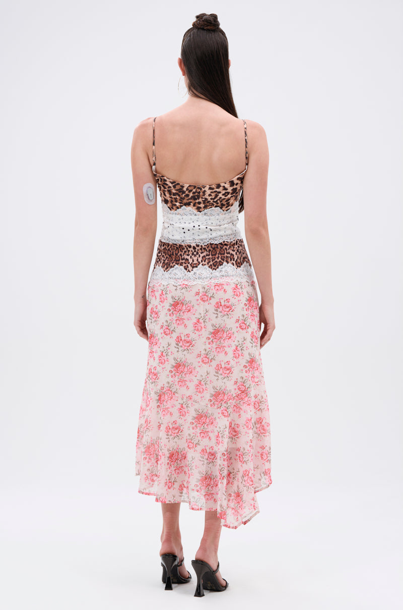 A woman with long dark hair stands with her back to the camera, wearing the CARRIE CODED SATIN MIDI DRESS, which features a leopard print top, white lace waist detail, and a pink floral chiffon skirt. She pairs it with black heels.