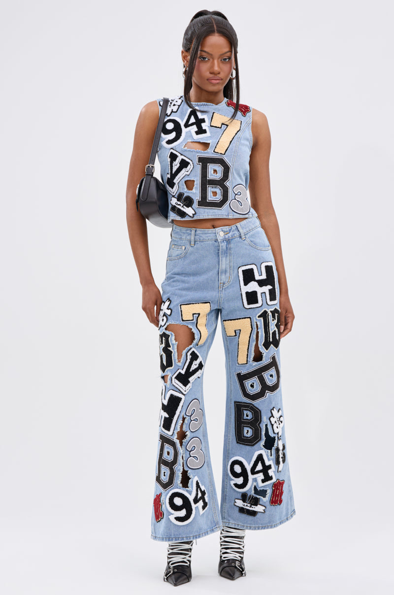 A woman stands facing forward in PATCH DENIM RELAXED PANT—wide-leg jeans with bold, varsity-inspired letter and number patches. She pairs them with a matching top, a small black shoulder bag, and black-and-white boots.