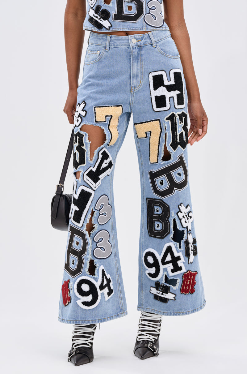 Someone wears the PATCH DENIM RELAXED PANT in light blue, featuring varsity-style number, letter, and cartoon patches, paired with a small black bag and pointed lace-up shoes.