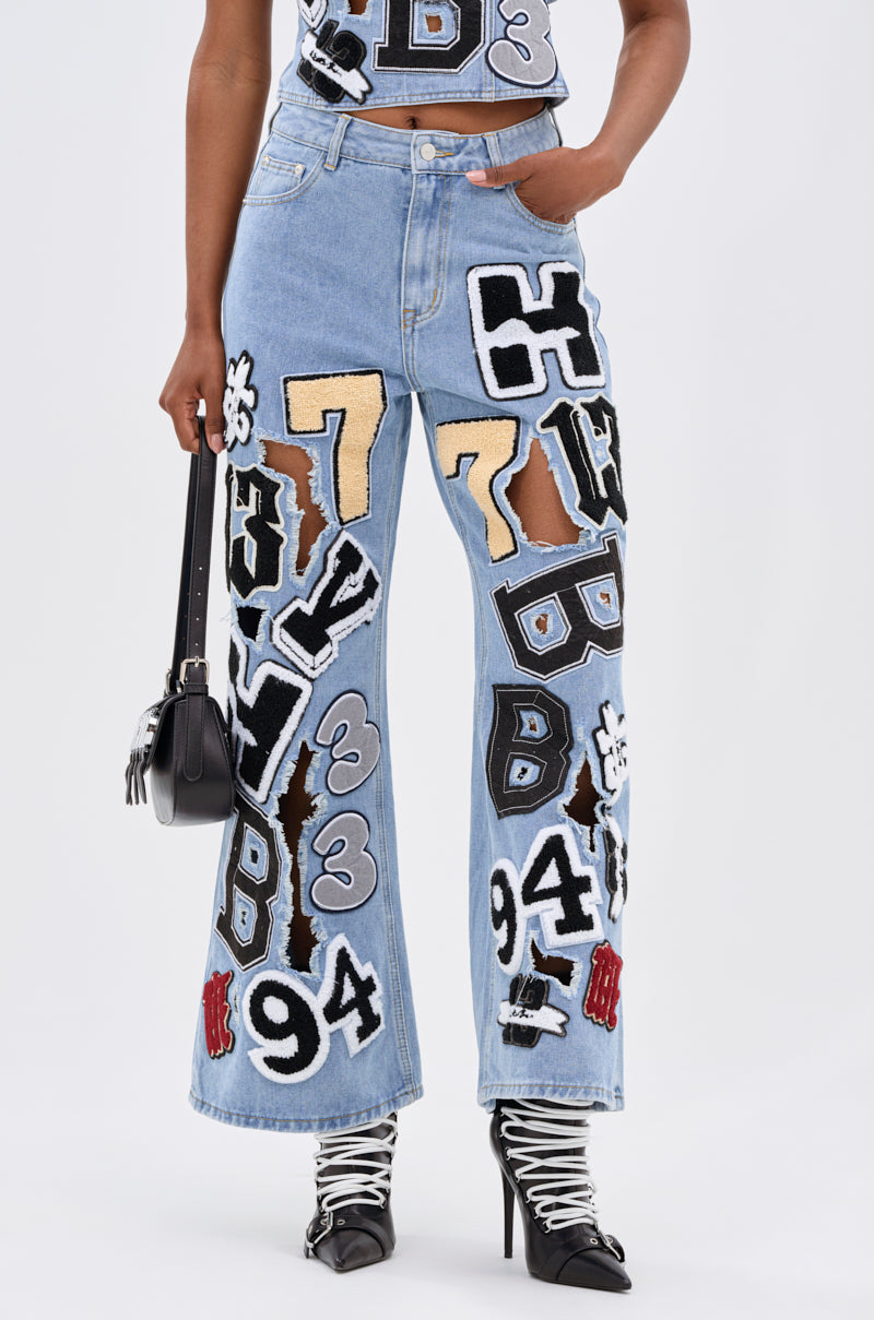 Wearing the PATCH DENIM RELAXED PANT—relaxed fit, light blue denim with large, colorful varsity-style number and letter patches—styled with black and white heeled boots and a small black handbag.