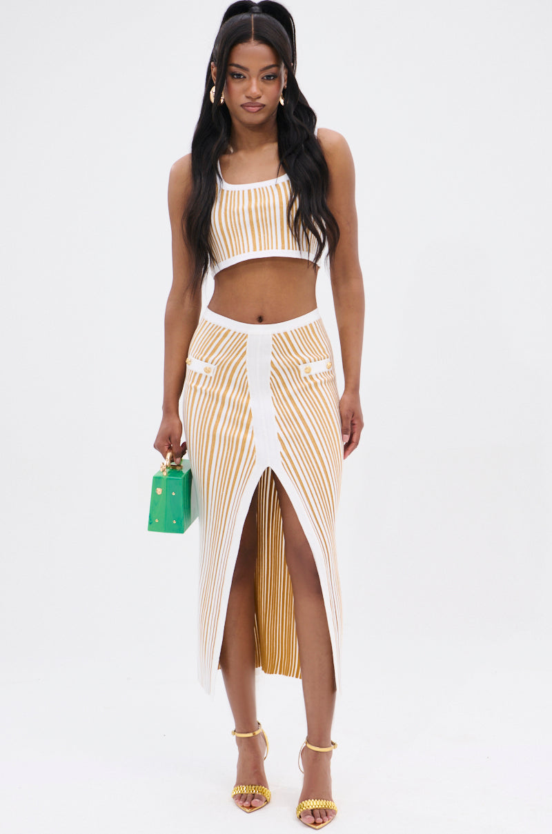 A woman confidently wears a striped white and yellow matching set with a sleeveless crop top and the LIFE IS GOOD MAXI SKIRT. The high-rise skirt, crafted from stretchy knit, features a high slit. She accessorizes with a small green handbag and yellow heels, while her wavy dark hair complements the look.