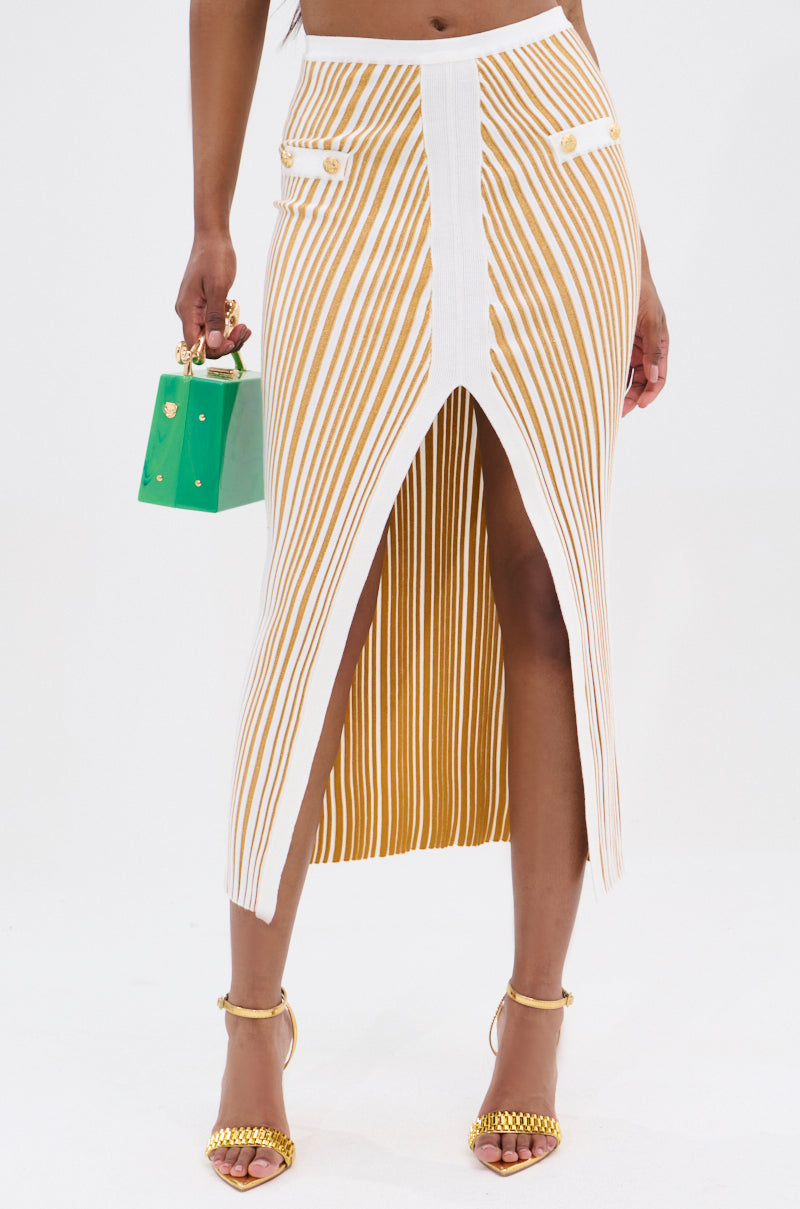 Wearing the LIFE IS GOOD MAXI SKIRT, showcasing a high-slit design in tan and white stripes made from stretchy knit fabric, paired with a small green handbag and yellow chain-detailed high-heeled sandals, this outfit shines against a plain white backdrop.