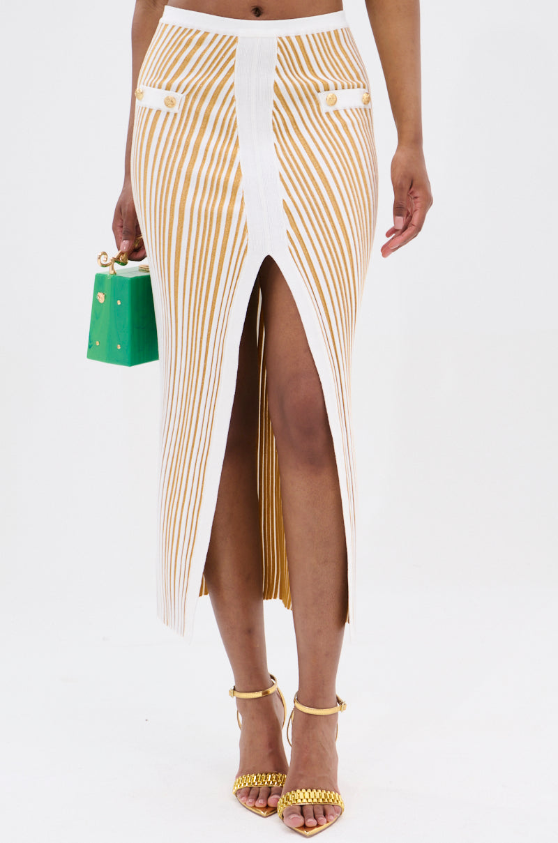 Dressed in the chic LIFE IS GOOD MAXI SKIRT featuring a white and mustard stripe and high-rise waist, they hold a small green handbag. Their gold strappy heels bring elegance to the plain white backdrop.