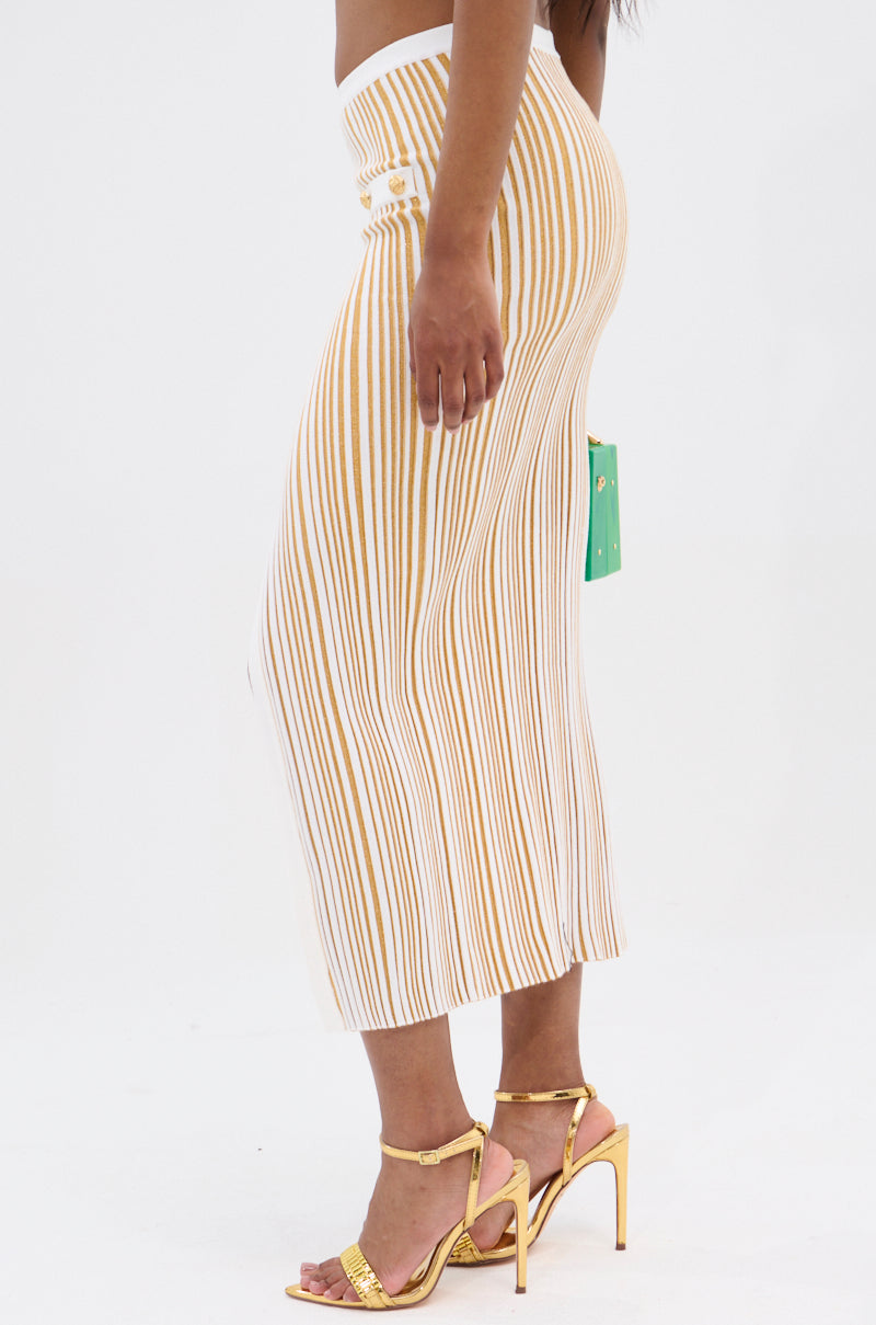 Wearing the LIFE IS GOOD MAXI SKIRT with white and gold stripes and a high-rise waist, this person pairs it with gold high-heeled sandals. They're holding a green rectangular bag against the plain white background.