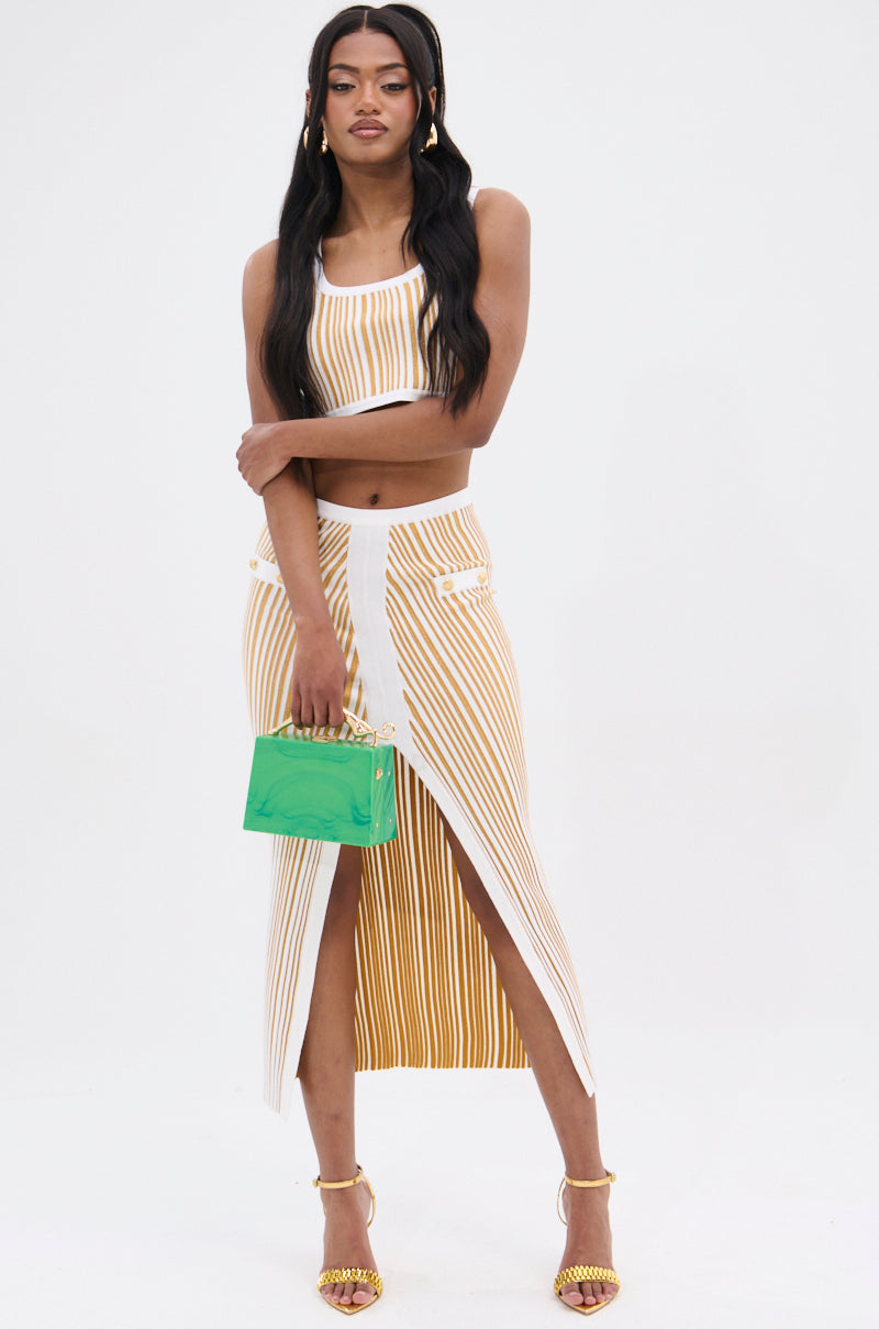 A person poses against a plain background in a striped crop top and the LIFE IS GOOD MAXI SKIRT, featuring a front slit. The high-rise waist and stretchy knit fabric offer a flattering fit. They carry a small green handbag, wear yellow high heels, and their long hair falls in loose waves.
