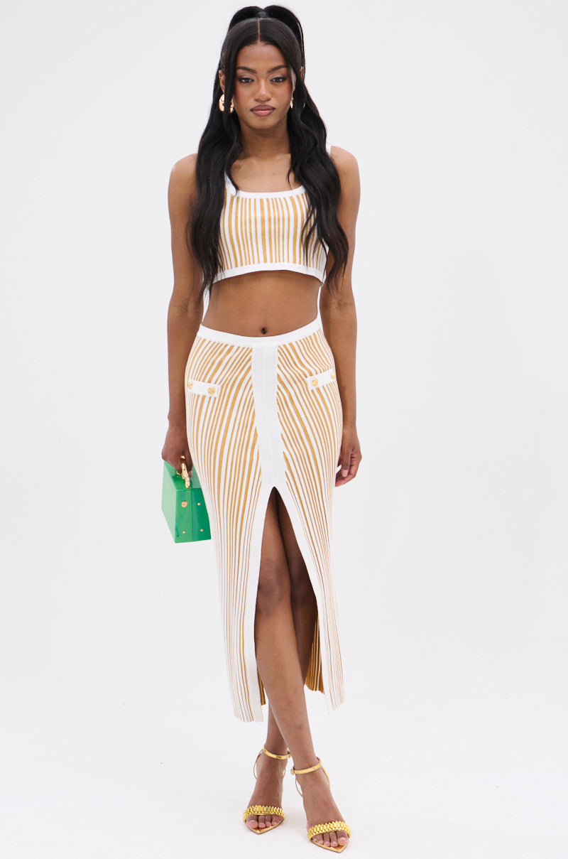 A woman in a striped crop top and the LIFE IS GOOD MAXI SKIRT with a front slit, paired with yellow heels. She carries a small green handbag featuring metallic accents and has long, wavy hair against a plain white background.