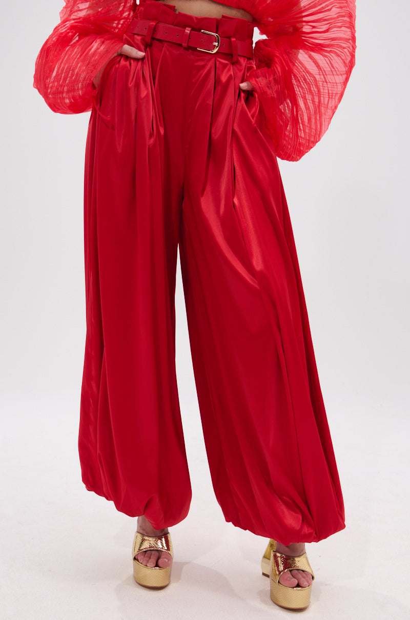 Wearing FORTUNE'S FAVORITE SATIN BELTED BALLOON PANT IN RED, styled with a sheer red top and gold platform sandals, a person poses against a plain white background.