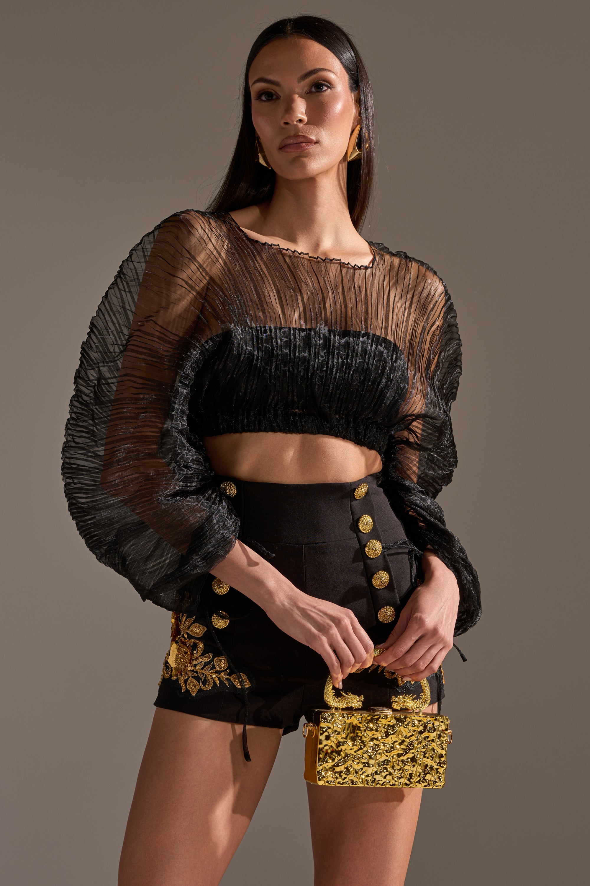 A woman poses confidently in the FORTUNES FAVORITE SHEER LONG SLEEVE CROP TOP with pleated details and voluminous sleeves, paired with high-waisted black shorts featuring gold buttons and embroidery, holding a small gold embellished handbag.