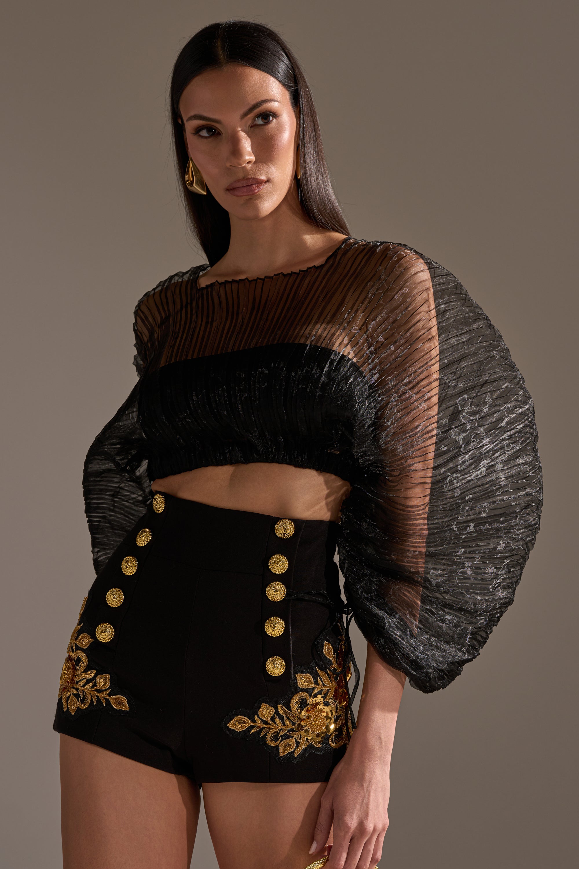 A woman with long dark hair models the FORTUNES FAVORITE SHEER LONG SLEEVE CROP TOP, featuring dramatic pleated sleeves, paired with high-waisted black shorts with gold buttons and floral embroidery, against a neutral background.