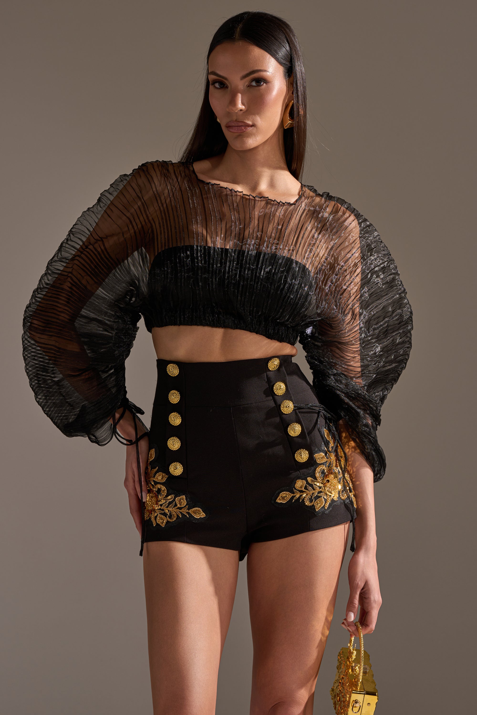 A woman poses in the FORTUNES FAVORITE SHEER LONG SLEEVE CROP TOP with pleated details and voluminous sleeves, styled with high-waisted black shorts featuring gold buttons, floral embroidery, and a small gold accessory.