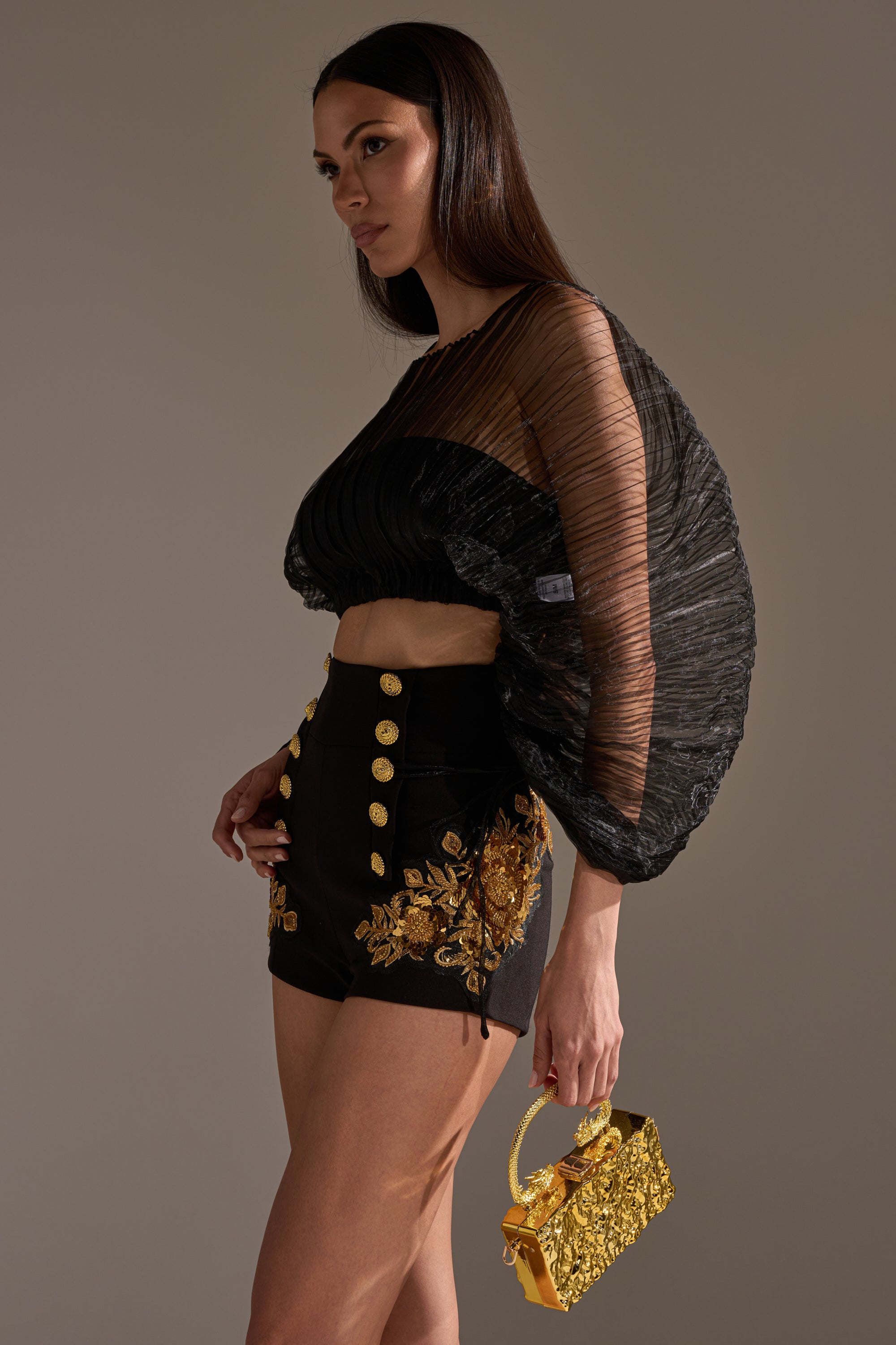 A woman poses in the FORTUNES FAVORITE SHEER LONG SLEEVE CROP TOP with pleated, voluminous sleeves, paired with black high-waisted shorts featuring gold embroidery and buttons, holding a small ornate gold handbag against a neutral background.
