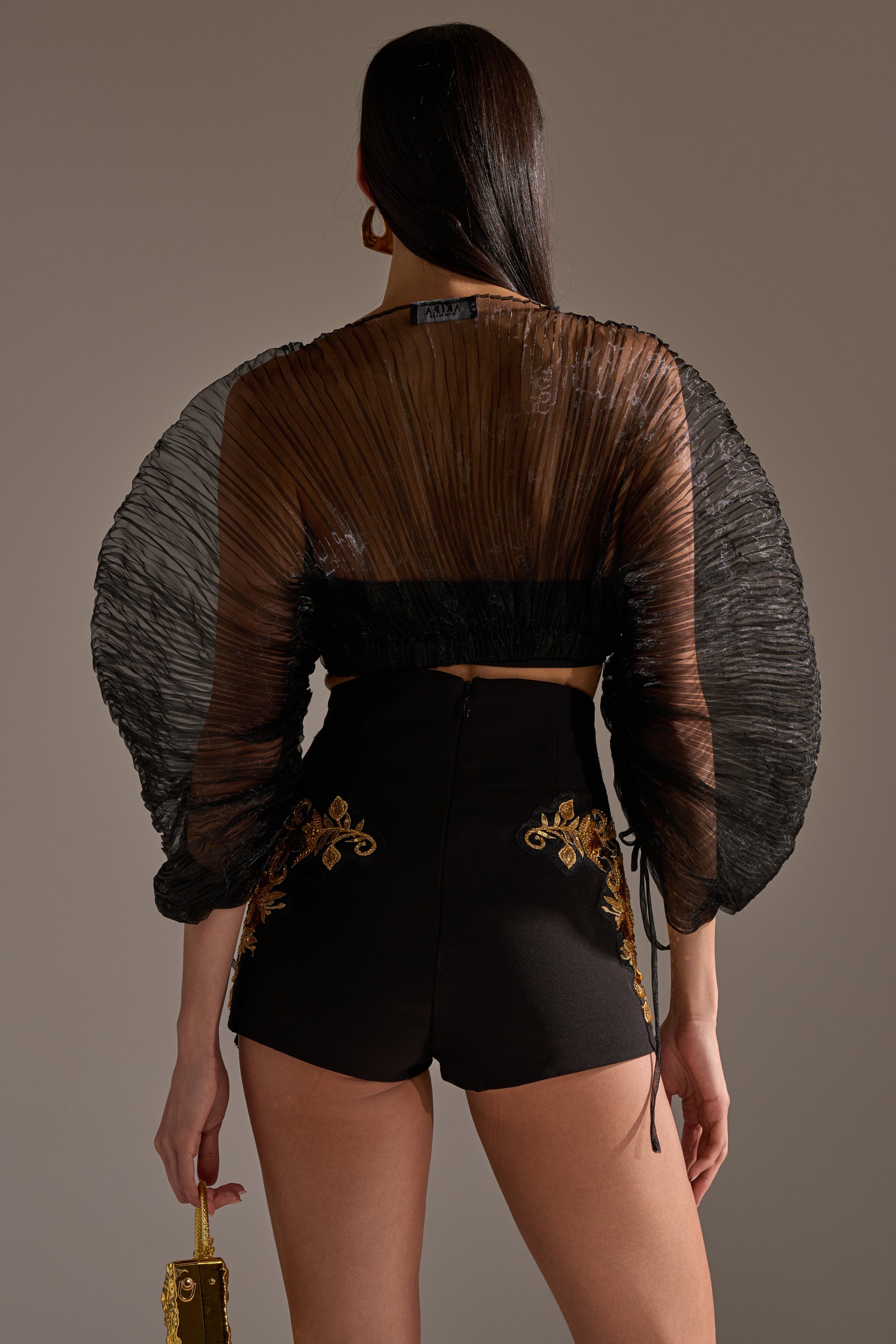 A woman stands facing away, wearing the FORTUNES FAVORITE SHEER LONG SLEEVE CROP TOP with pleated details and voluminous sleeves. She pairs it with high-waisted black shorts with gold floral embroidery and holds a small gold handbag in her left hand.