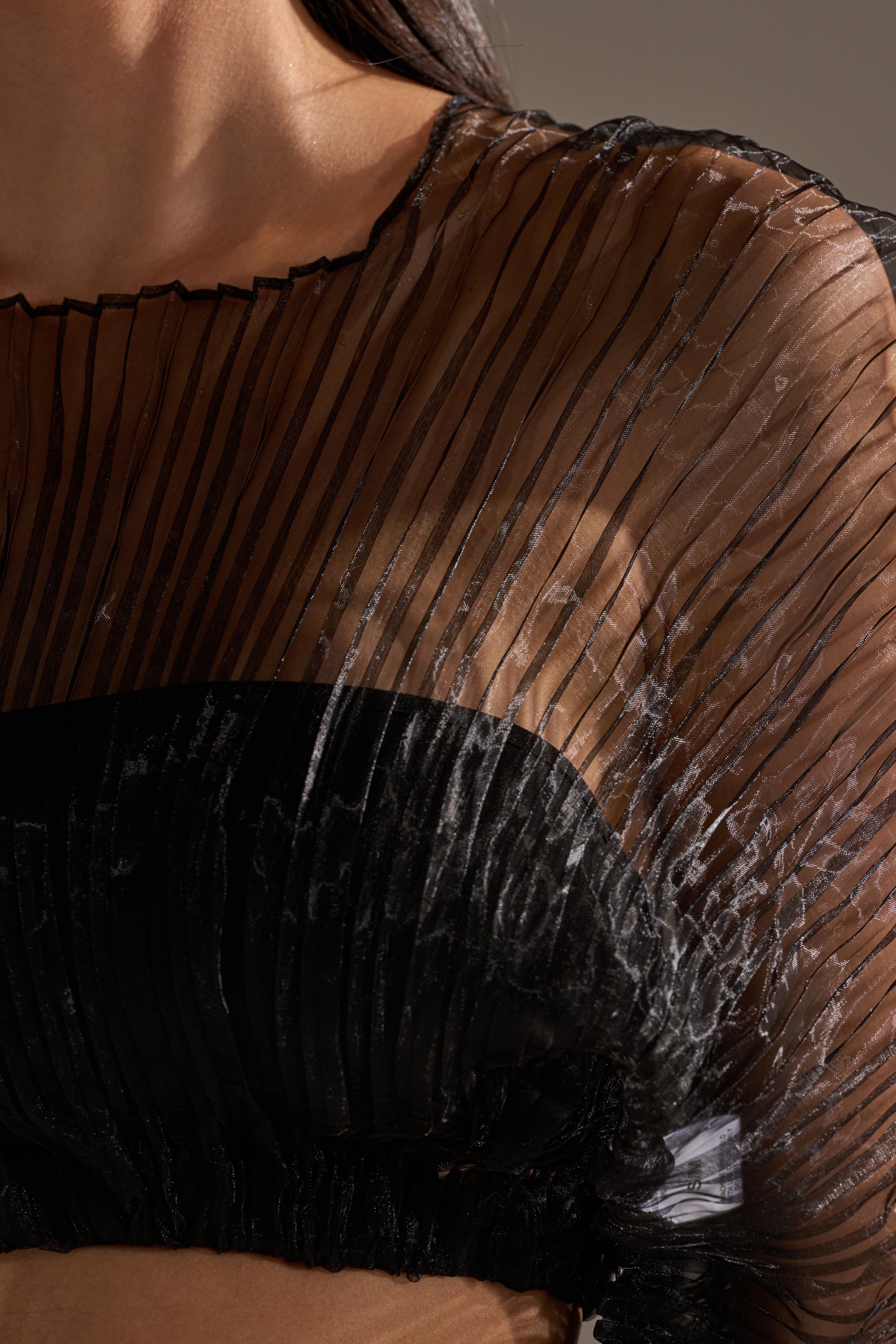 A close-up shows someone wearing the FORTUNES FAVORITE SHEER LONG SLEEVE CROP TOP with pleated, transparent fabric over the shoulder and chest, partially revealing a black undergarment underneath.