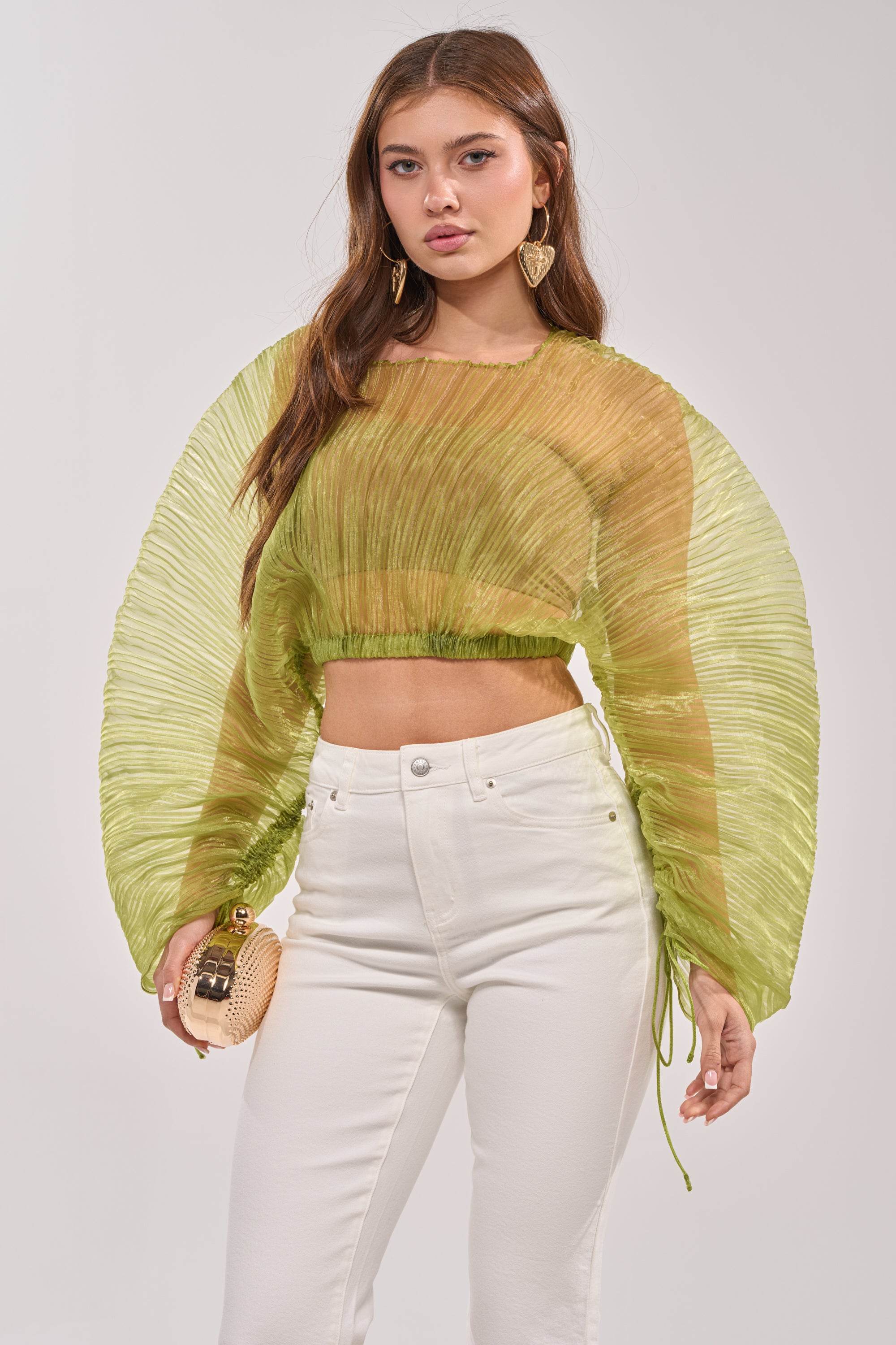A woman wears the BEYOND PRETTY PLEATED COCOON BLOUSE IN GREEN, paired with white high-waisted pants and a small round beige clutch. She has long brown hair, statement earrings, and a confident expression. 0