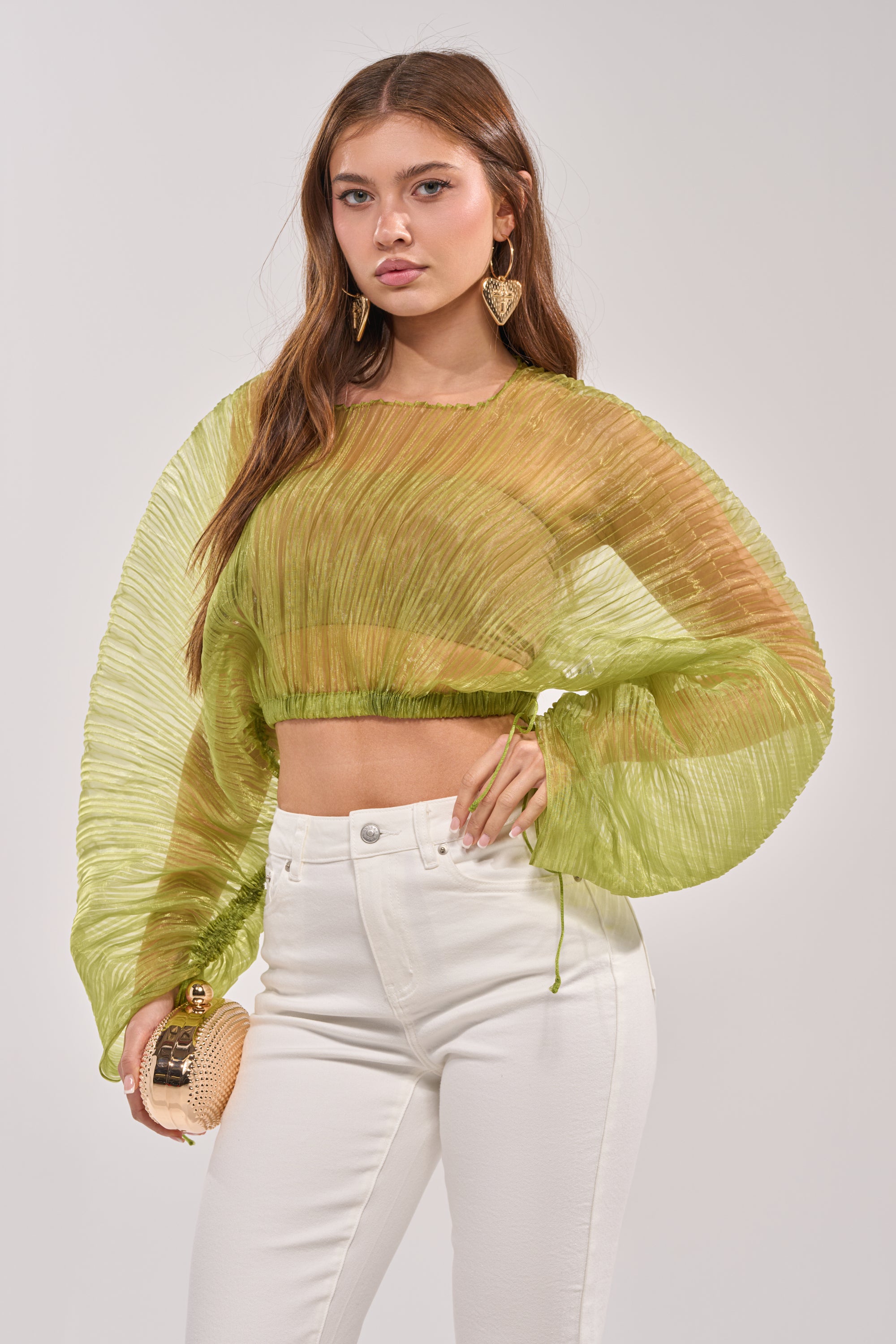 A woman wears the BEYOND PRETTY PLEATED COCOON BLOUSE IN GREEN with high-waisted white pants and holds a small round clutch while posing against a plain light-grey background. 1