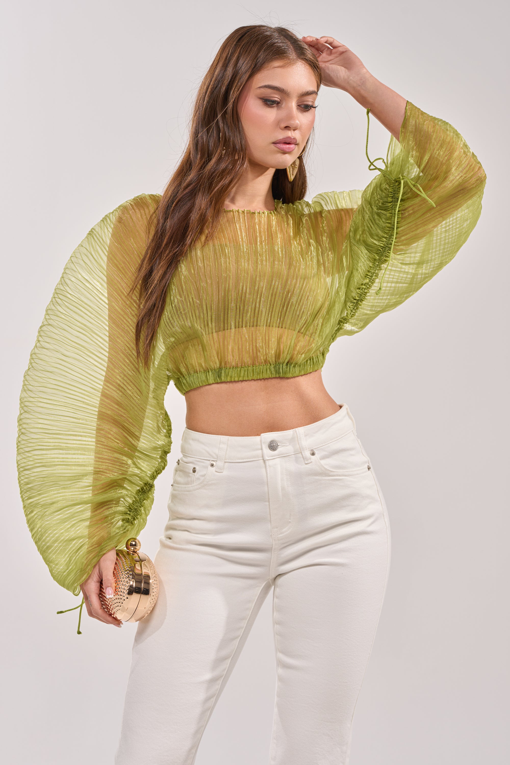 A woman in the BEYOND PRETTY PLEATED COCOON BLOUSE IN GREEN and high-waisted white pants holds a small round beige purse, posing with one hand on her head against a plain background. 2