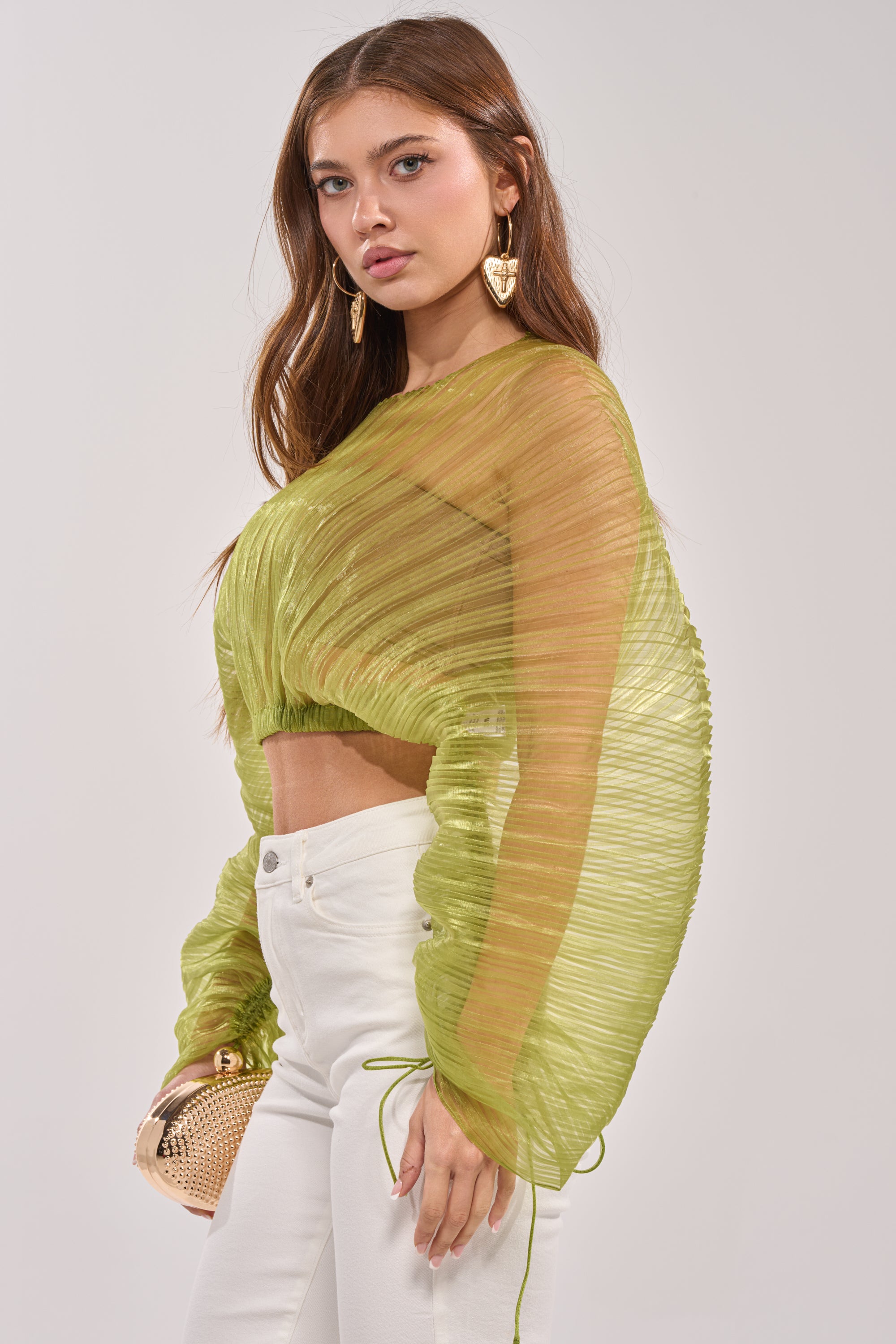 A woman wears the BEYOND PRETTY PLEATED COCOON BLOUSE IN GREEN with white high-waisted pants, accessorized with a small gold clutch and gold heart-shaped earrings. She has long brown hair and a neutral expression. 3