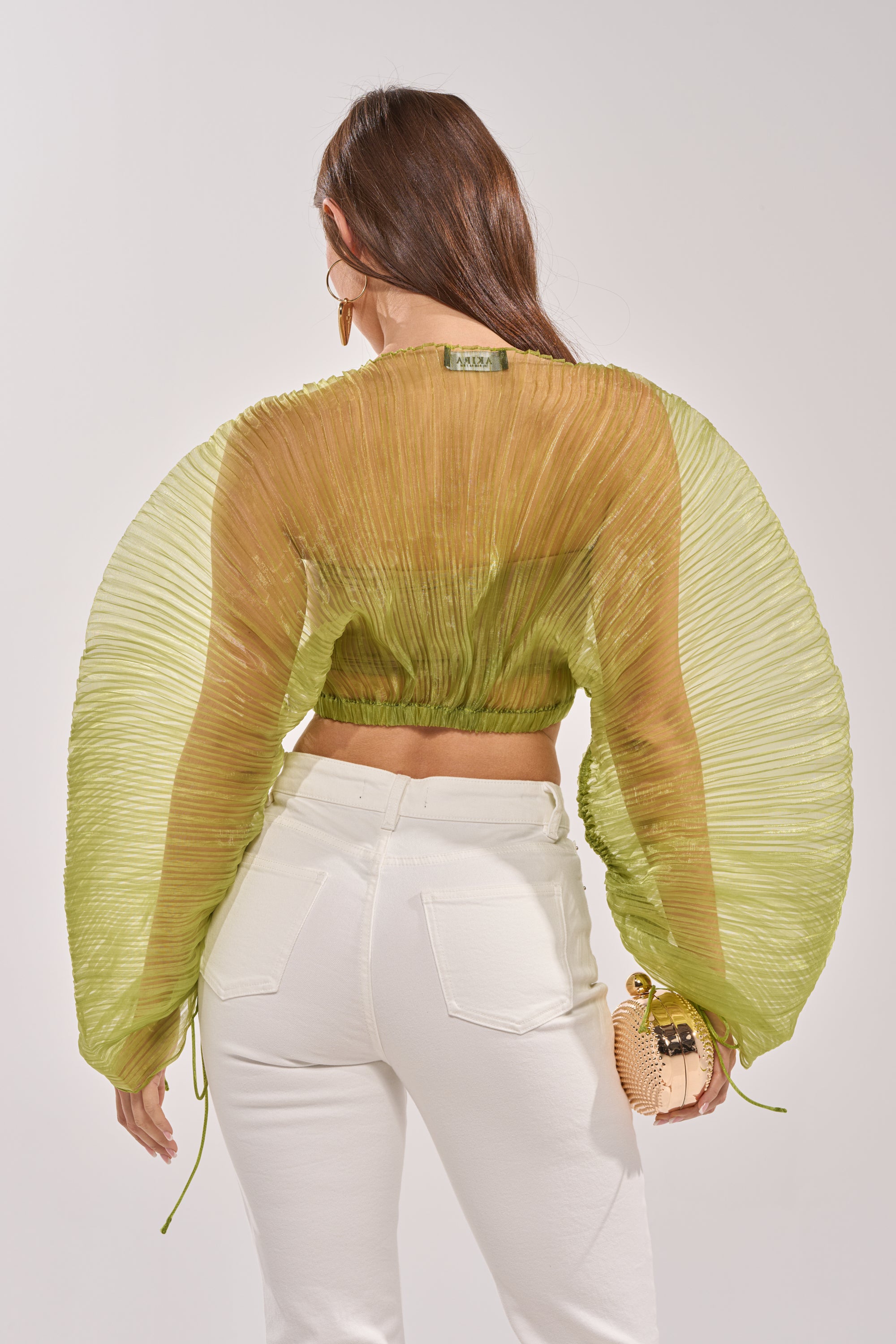 A woman with long brown hair stands facing away, wearing the BEYOND PRETTY PLEATED COCOON BLOUSE IN GREEN—a sheer, pleated chiffon top with voluminous sleeves—paired with high-waisted white jeans and holding a textured beige clutch. 4