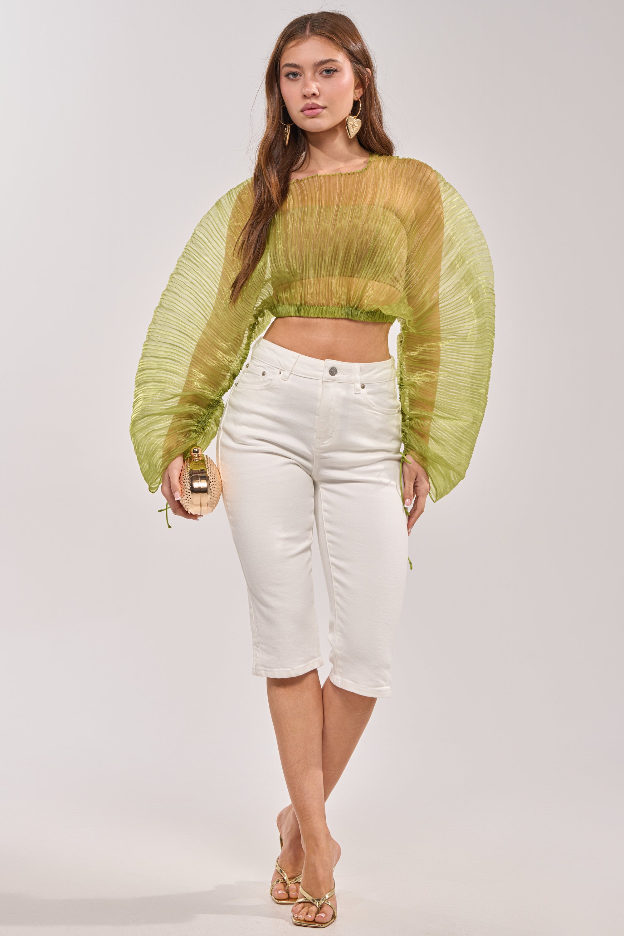 A woman poses in the BEYOND PRETTY PLEATED COCOON BLOUSE IN GREEN with pleated long sleeves and high-waisted white cropped pants, accessorized with a small purse, gold earrings, and heels against a light background. 5