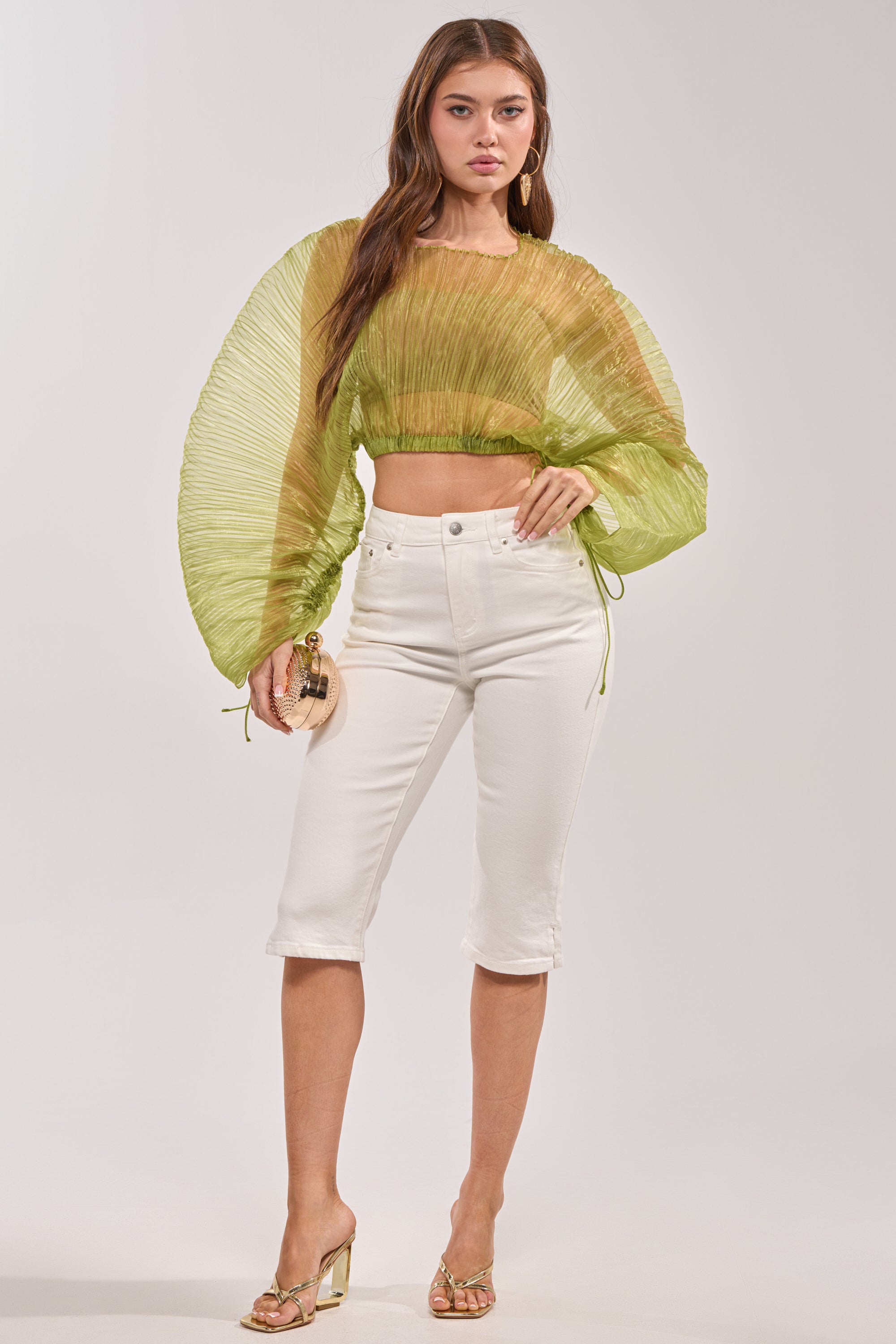 A woman wears the BEYOND PRETTY PLEATED COCOON BLOUSE IN GREEN with white fitted knee-length pants, clear-heeled sandals, and holds a small round clutch. 6