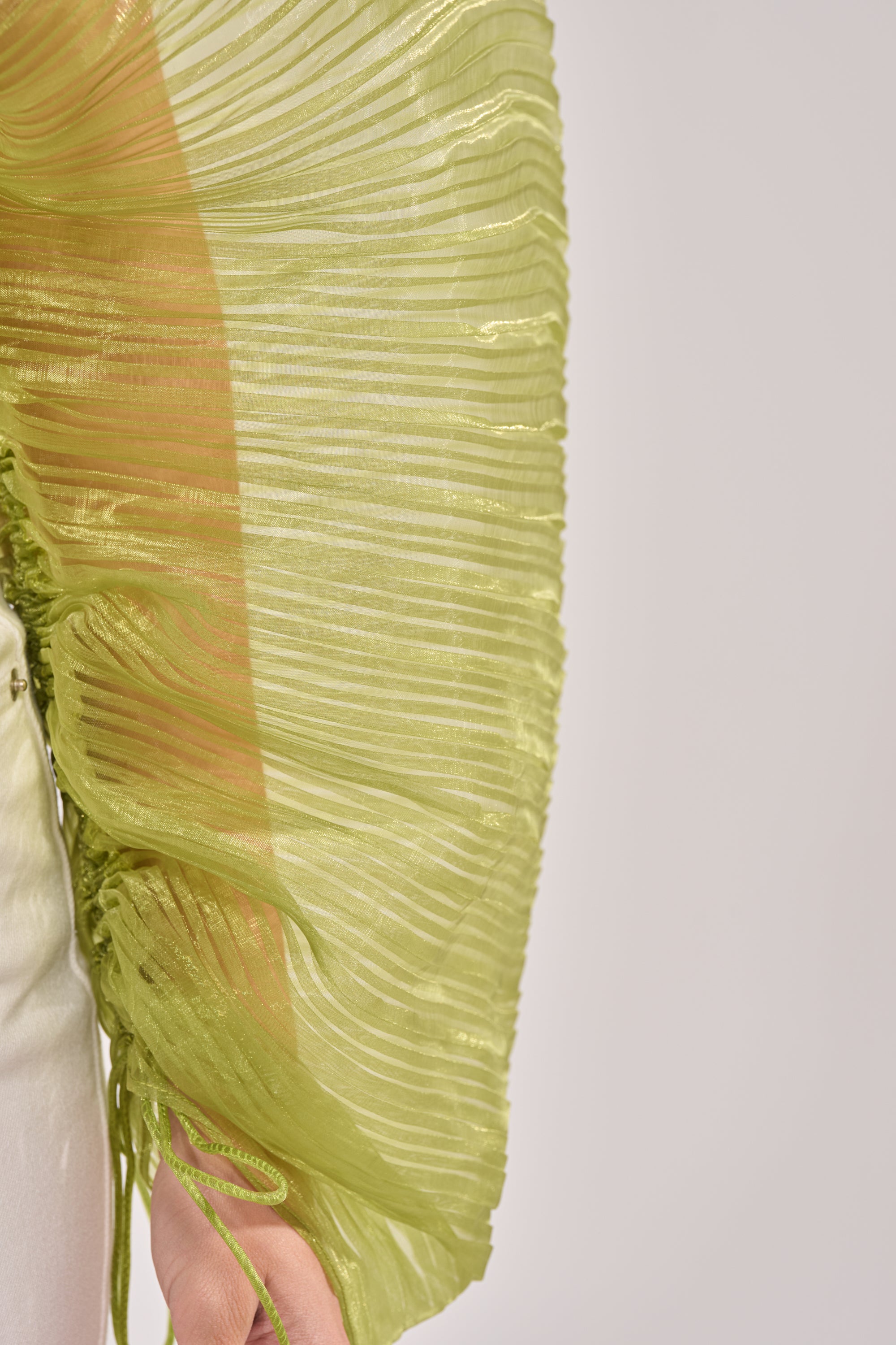 A close-up of the BEYOND PRETTY PLEATED COCOON BLOUSE IN GREEN, featuring sheer chiffon fabric with pleats and a ruffled texture draped over an arm, set against a light background. The blouse’s delicate material appears partially transparent. 7