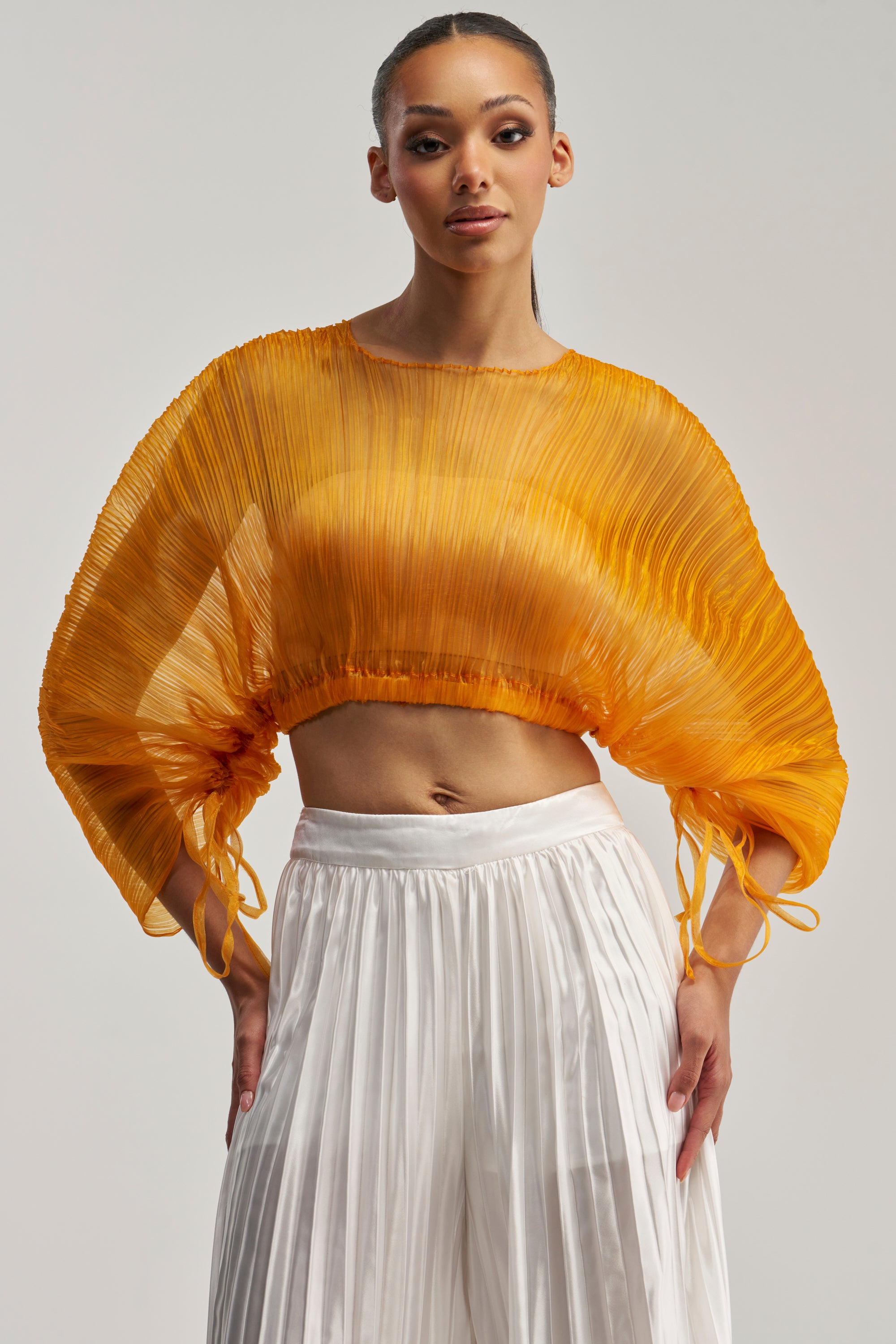 Wearing the FORTUNES FAVORITE SHEER LONG SLEEVE BLOUSE IN MARIGOLD, a woman poses against a plain light background, paired with white, high-waisted, wide-leg pleated pants.