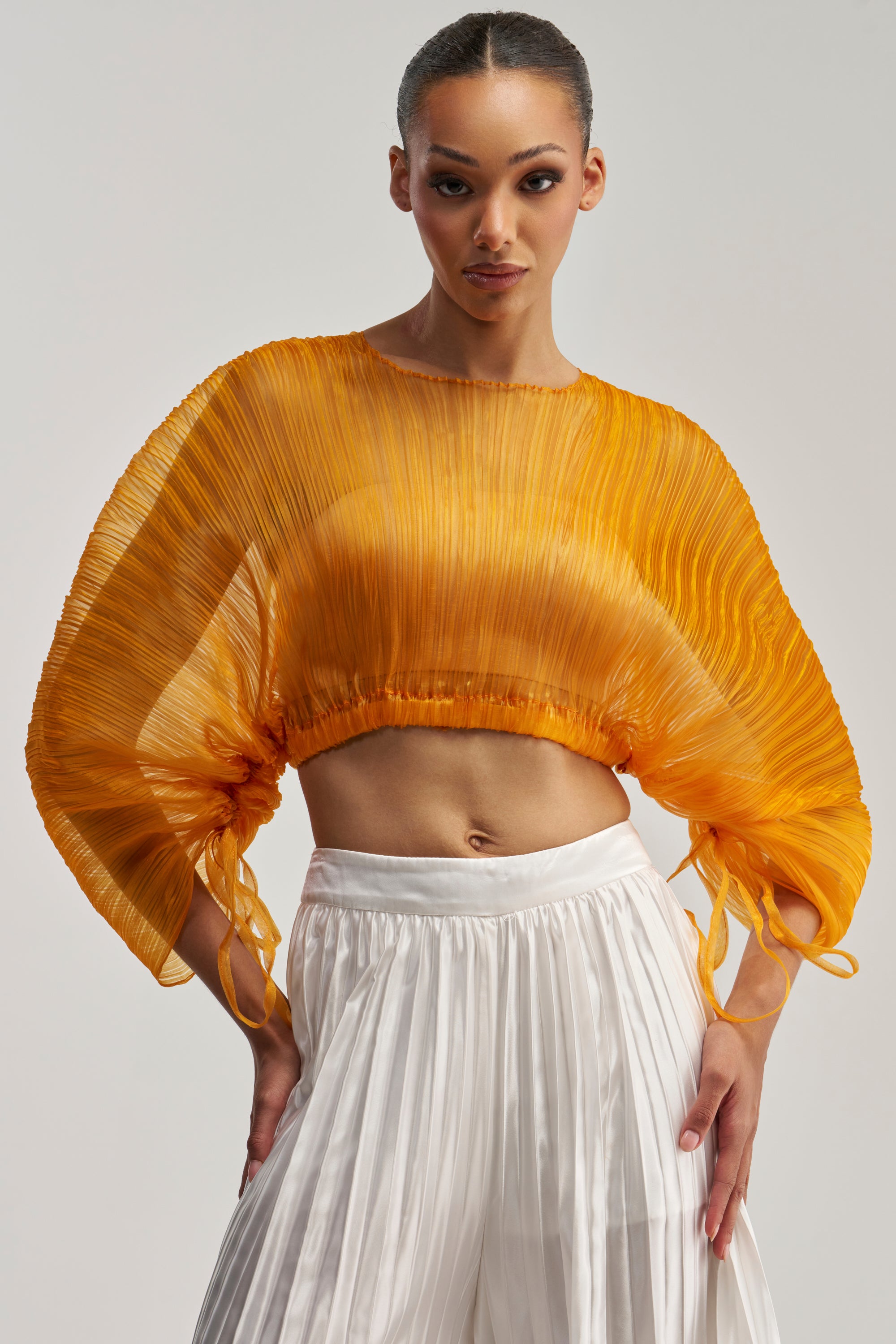 A woman wearing the FORTUNES FAVORITE SHEER LONG SLEEVE BLOUSE IN MARIGOLD—featuring pleated detailing and wide, sheer long sleeves—is styled as a chic going-out top, paired with high-waisted white pleated pants and standing against a plain background.