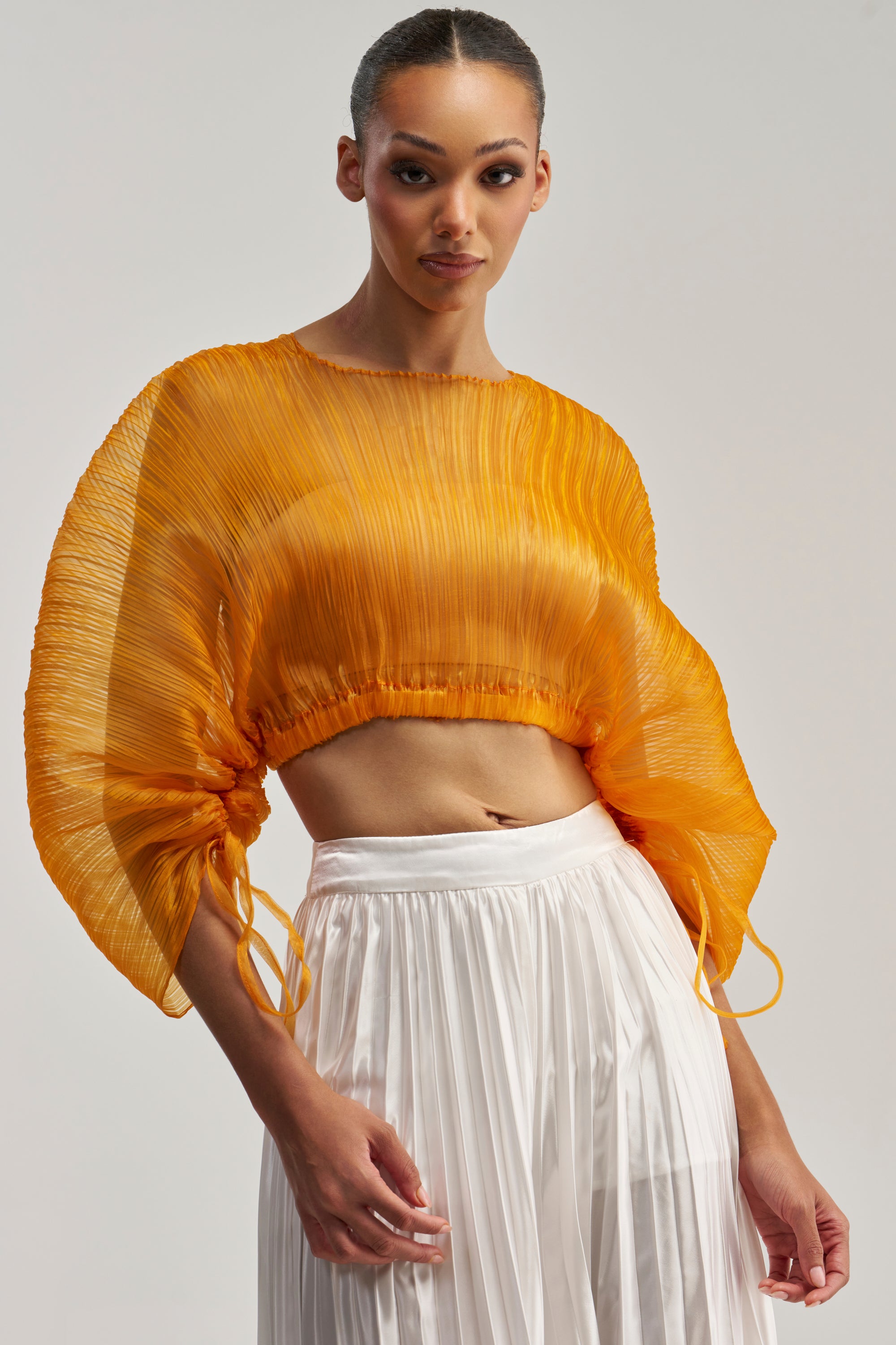 Wearing the Fortunes Favorite Sheer Long Sleeve Blouse in marigold, a woman poses against a neutral background. The blouse features pleated details and voluminous sleeves, paired with a high-waisted, white pleated skirt.